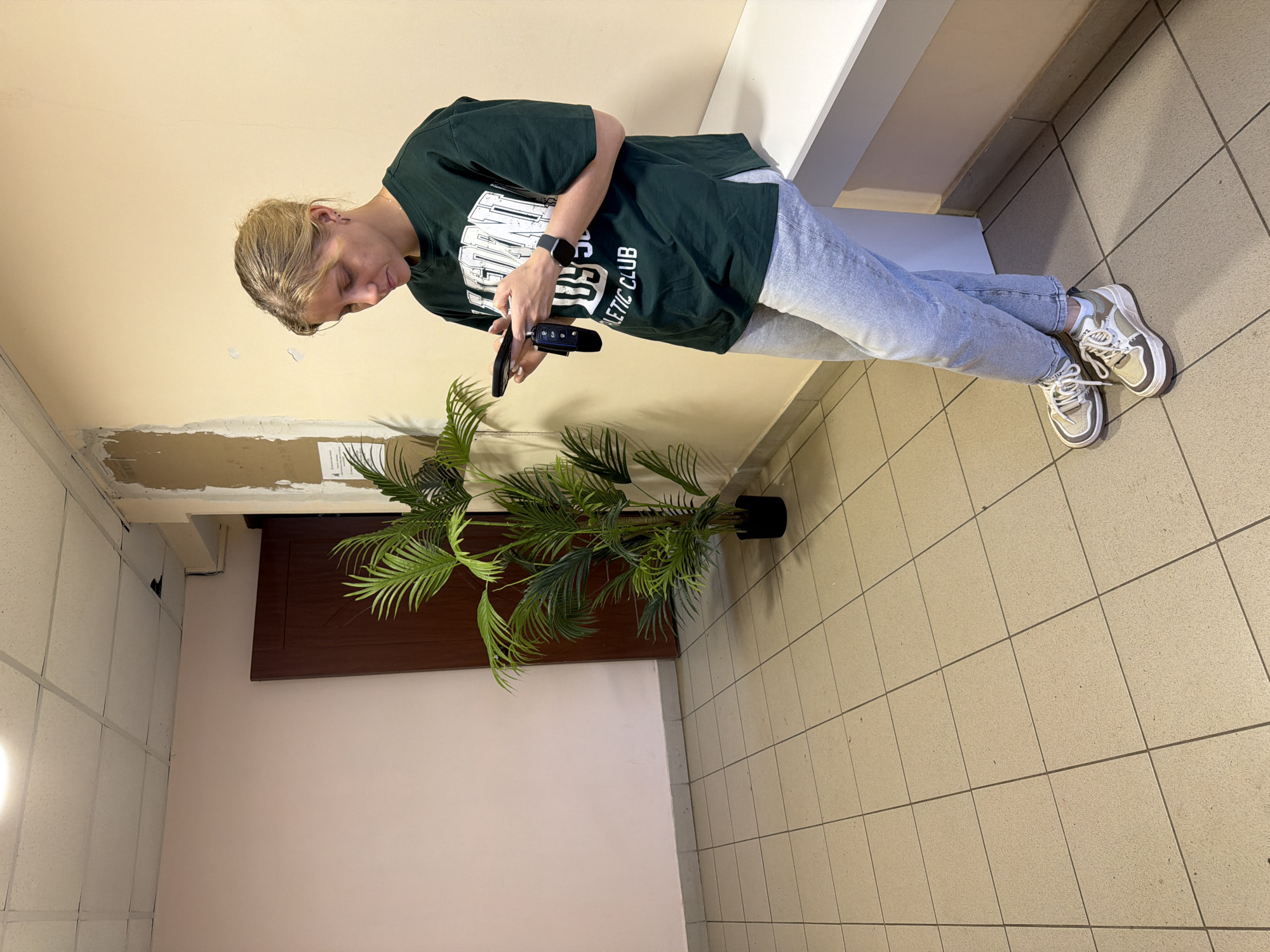 A young woman stands indoors on a tiled floor near a large potted plant. She is wearing a dark green t-shirt with white lettering, light blue jeans, and off-white sneakers. She is looking down at something in her hands, possibly a phone or wallet. The background consists of beige walls, a white ledge, and a closed wooden door. The location is likely Chaikovsky, Russia.