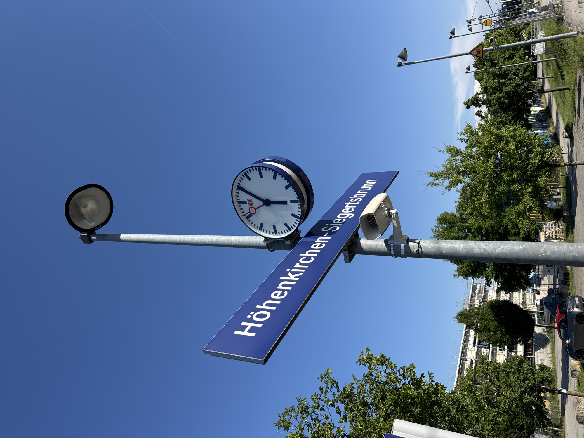 An outdoor, eye-level, medium shot captures a sign for the train station in Höhenkirchen-Siegertsbrunn, Germany, under a clear blue sky. The blue sign with white lettering reads "Höhenkirchen-Siegertsbrunn". Attached to the same pole as the sign is a large, white, round clock with blue markings and a logo that reads "DB". A street lamp is visible above the clock and sign. In the background, trees line a sidewalk, and a building is under construction, with scaffolding visible. A small, red-topped car is parked on the side of the road. Several other street lamps and warning signs are visible further down the street.