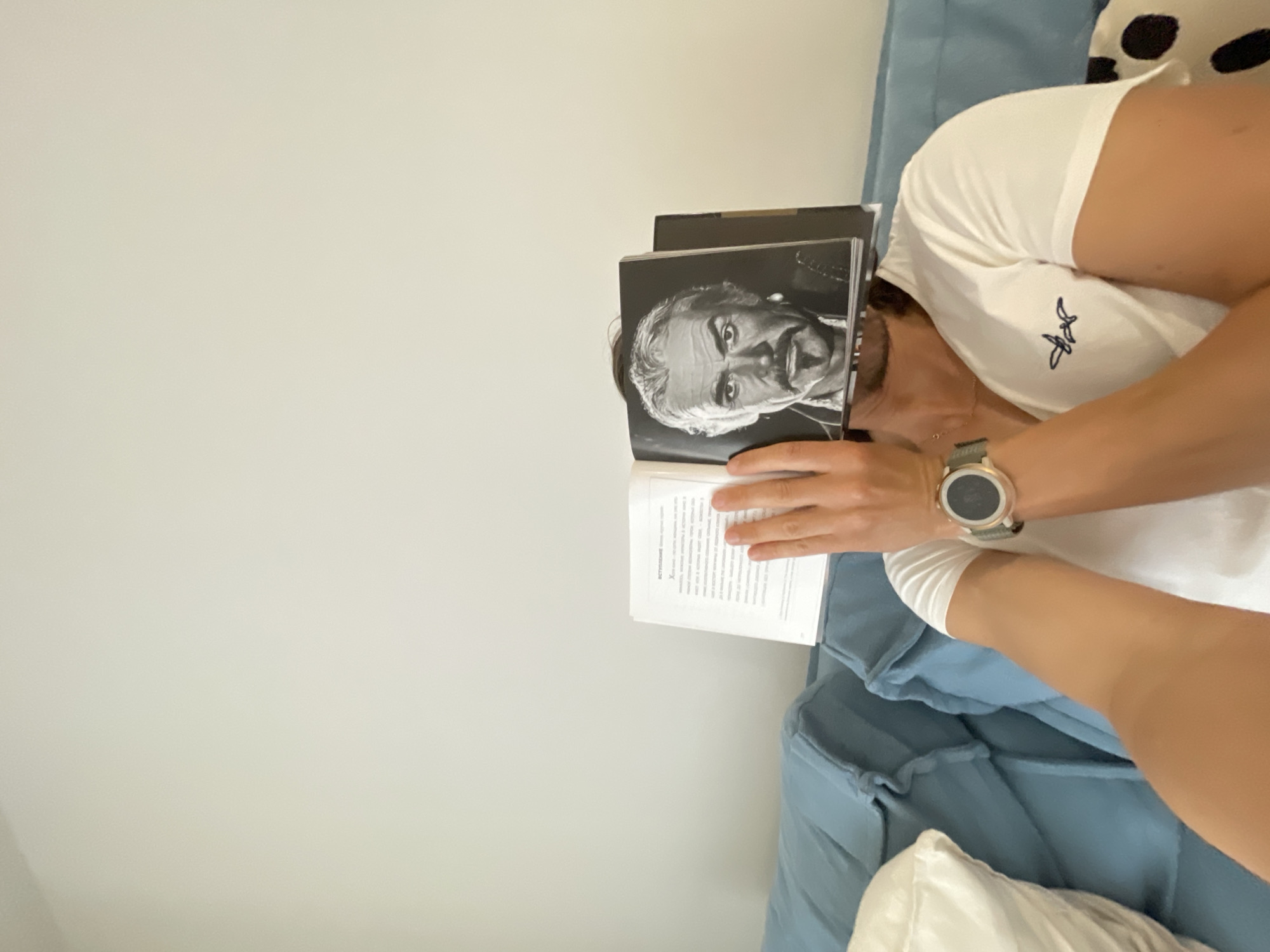 The image shows a person reclining on a blue couch. They are wearing a white t-shirt and a watch. The person is holding an open book in front of their face, obscuring it. The cover of the book features a black-and-white portrait of a man. The book appears to be being read. The environment consists of a white wall and a light blue couch.