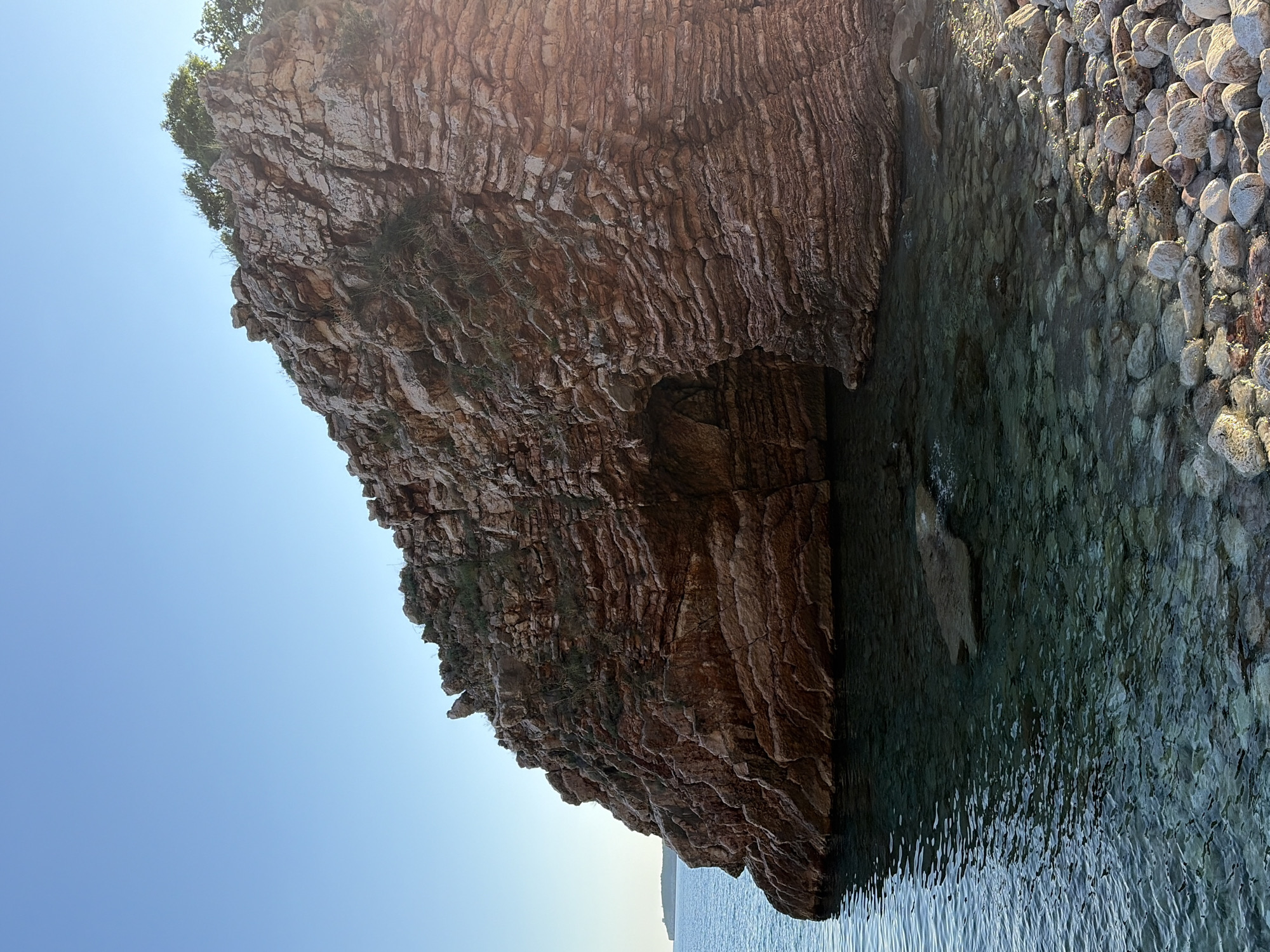 A large, layered rock formation extends into the water. A small cave is visible at the base of the rock. The water is clear and reflects the rock. The sky is a light blue. The right side of the rock formation is covered in rounded pebbles. Based on the coastal geology and vegetation, the location is likely in the area of Rafailovići, Montenegro.