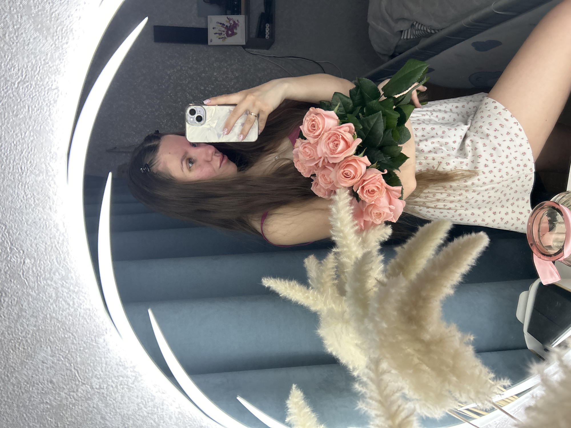 A person is taking a selfie in a mirror, holding a bouquet of pink roses. The individual is wearing a white dress with small red floral patterns. A smartphone is visible in their hand, and there are other items in the background, including pampas grass in the mirror. The location appears to be indoors, possibly in a room with a blue upholstered wall visible in the mirror. I am unable to determine if this image was taken in Klin, Russia.