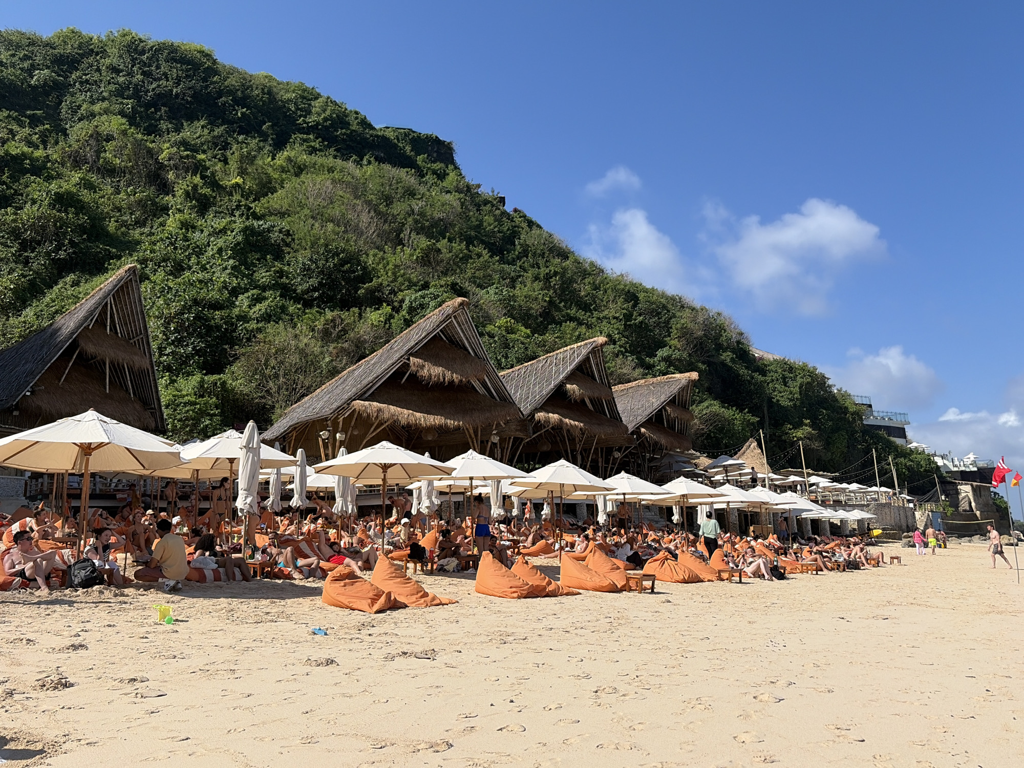 The image shows a crowded beach scene on a sunny day in Kuta Selatan, Indonesia. In the foreground, there is a wide expanse of sandy beach with numerous orange beanbag chairs and white umbrellas providing shade for beachgoers. Many people are seated on the beanbags, some appear to be lounging, others are conversing, and some are looking out towards the ocean. In the midground, there are several thatched-roof structures, resembling huts or cabanas, set back from the beach. Behind these structures, a lush, green hillside rises steeply. The sky is clear and blue with a few scattered white clouds. The overall impression is a vibrant and popular tourist destination.