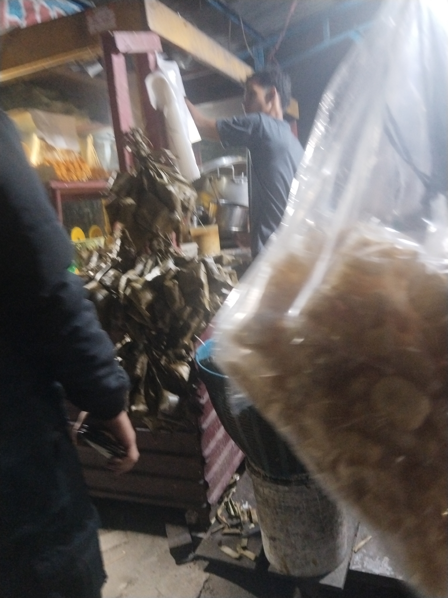 The image depicts a dimly lit scene at what appears to be a street food stall in Muara Bulian, Indonesia. In the foreground, a person wearing a dark garment is partially visible on the left. To the right, a large, transparent plastic bag filled with what seems to be dried food items or ingredients is prominently displayed. In the background, another individual wearing a dark t-shirt is seen standing and possibly interacting with the stall. The stall itself has wooden structures and shelves displaying various items, including what looks like food wrapped in leaves, and some metal containers. A blue bucket is also visible near the base of the stall. The overall impression is one of a busy, informal marketplace or food vendor setup.