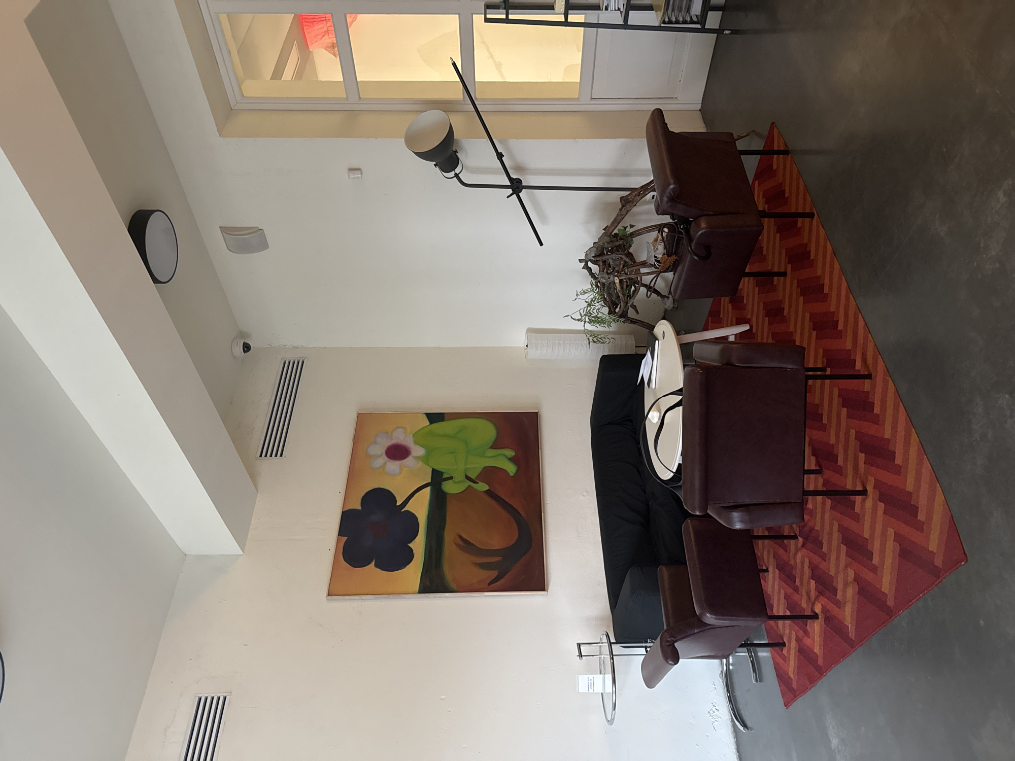 The image depicts a seating area in a room with white walls. A painting of two flowers, one white and one dark blue, hangs on the wall next to a black sofa. Two leather armchairs are placed on a red, orange, and brown herringbone patterned rug. A black floor lamp stands near the armchairs, and a small round table with a white top is between the armchairs and the sofa. The room has a modern, minimalist aesthetic with a combination of natural and artificial light. The location is not possible to determine based solely on visual data.