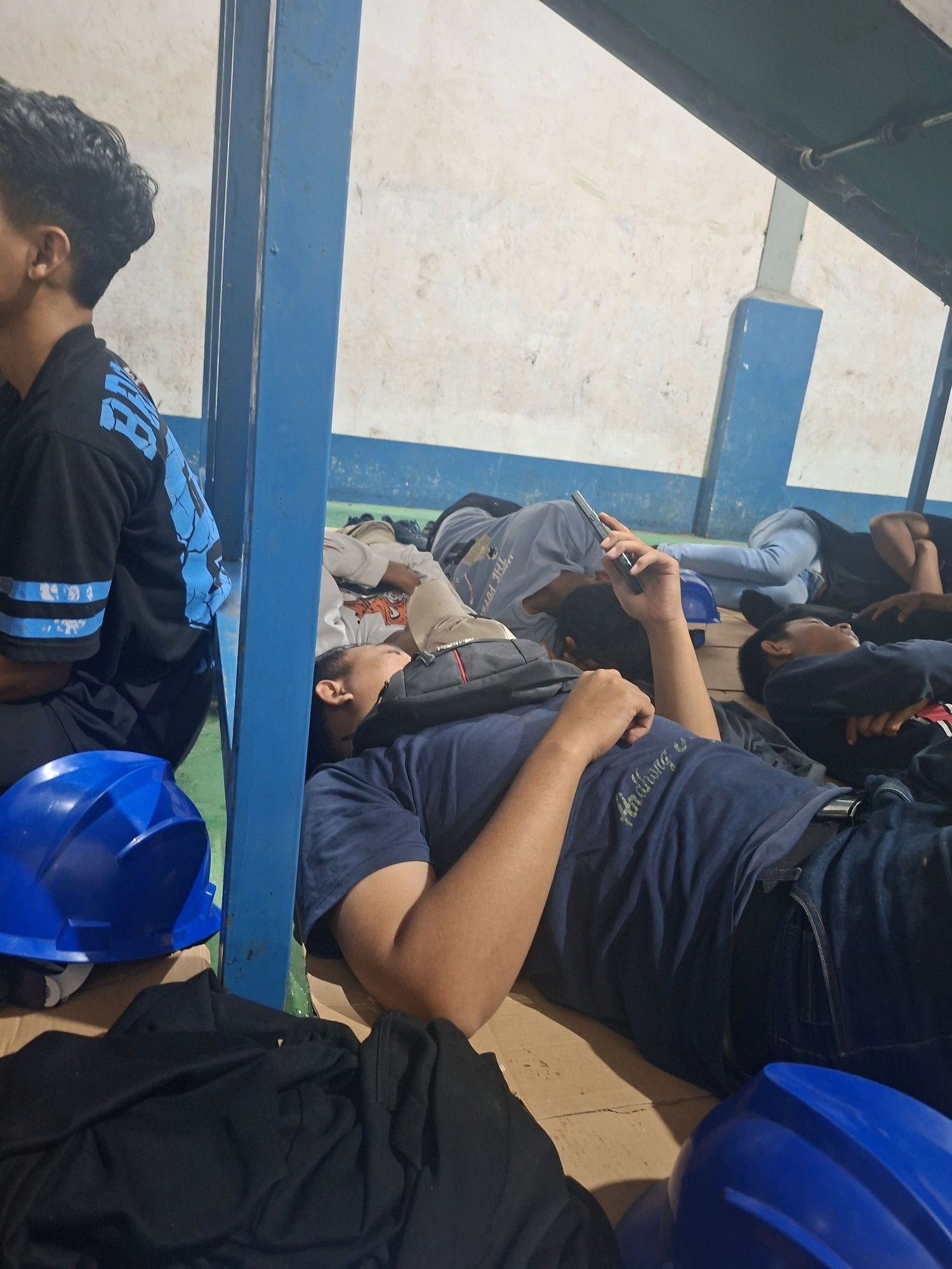 The image depicts several individuals, appearing to be young men, lying down on a floor, possibly resting or sleeping. They are situated indoors, in what seems to be a communal space with a textured, light-colored wall and a blue support structure. The individuals are casually dressed, some in t-shirts and jeans. One person in the foreground is holding a mobile phone. Blue hard hats are visible on the floor in the foreground, suggesting a work-related context. The overall impression is one of rest or downtime within a functional, possibly industrial or construction-related, environment. The specific location in Ngimbang, Indonesia, is not visually evident from the image itself, but the context suggests a utilitarian setting where such hard hats might be used.