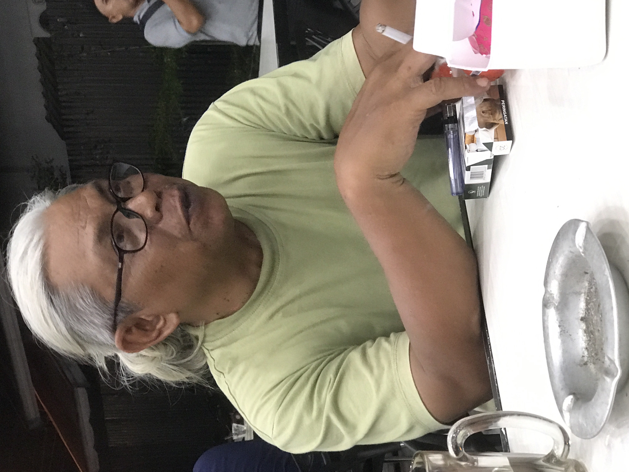An elderly man with gray hair and glasses is seated at a white table, holding a lit cigarette. He is wearing a pale green t-shirt. On the table, there is a pack of cigarettes, a lighter, an ashtray, and a clear glass mug. Another individual wearing gray is partially visible in the background, out of focus. The photo appears to be taken in Lhokseumawe, Indonesia.