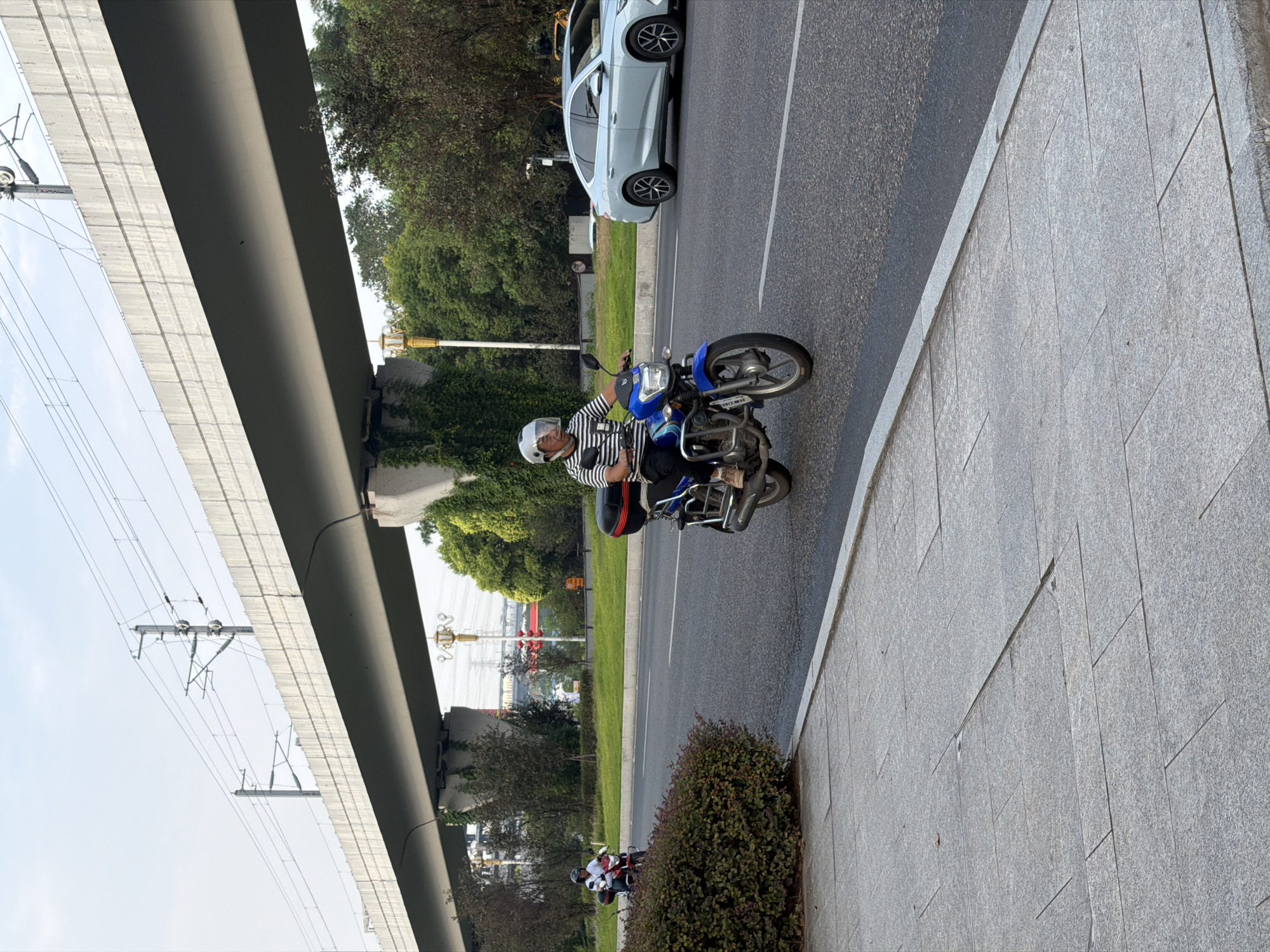 In Jiangjin City, China, a man wearing a white helmet and a striped shirt rides a blue motorcycle on an asphalt road. He is positioned in the foreground, centered on the road, with a silver car to his right, traveling in the same direction. To the left of the road, a dense green bush lines the concrete sidewalk. In the background, an elevated concrete structure, likely part of a transportation system, spans across the scene, supported by concrete pillars. Electrical wires and poles are visible above the elevated structure, indicating its function. Green trees and foliage surround the base of the structure and line the median of the road. The sky is overcast, suggesting either early morning or late afternoon.