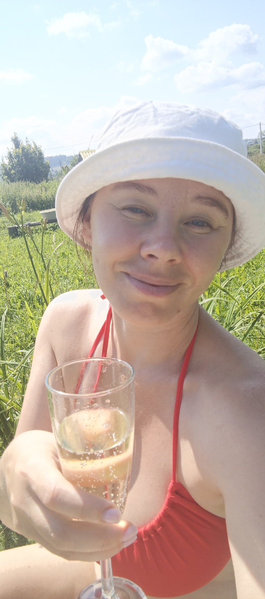 A selfie of a woman wearing a white bucket hat and a red bikini top, holding a glass of champagne in her right hand, is taken outdoors on a sunny day. The woman is smiling and looking towards the camera, with her eyes partially closed. Her skin appears to be slightly tanned, and she has light-colored eyes and eyebrows. The background consists of lush green grass and some plants, with a blurred background of trees and a structure that may be a building. The sky is visible, with scattered white clouds. The overall impression is a casual and relaxed moment of enjoyment in a natural setting. The location is not specified in the image, but it is described as being in the city of Bolshoy Bukor, Russia.