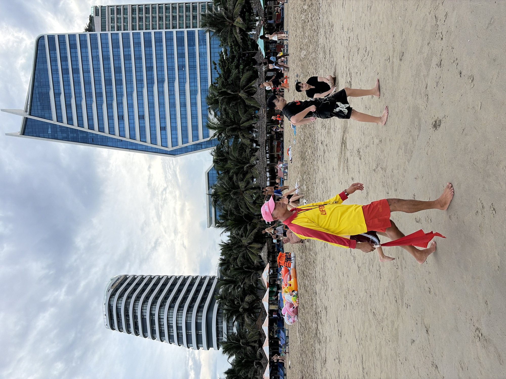 The image depicts a beach scene in Đà Nẵng, Vietnam, with two modern high-rise buildings in the background. In the foreground, a lifeguard wearing a bright yellow and red shirt with "LIFEGUARD" printed on it and a pink cap walks across the sand, carrying a red flag. To his right, a young person in black shorts and t-shirt walks away from the camera, and further to the right, another person sits on the sand. The beach is dotted with sun loungers, umbrellas, and inflatable toys, suggesting a day with beachgoers. Palm trees line the area behind the beach loungers, and the sky is overcast with clouds. The modern architecture of the buildings contrasts with the natural beach environment.