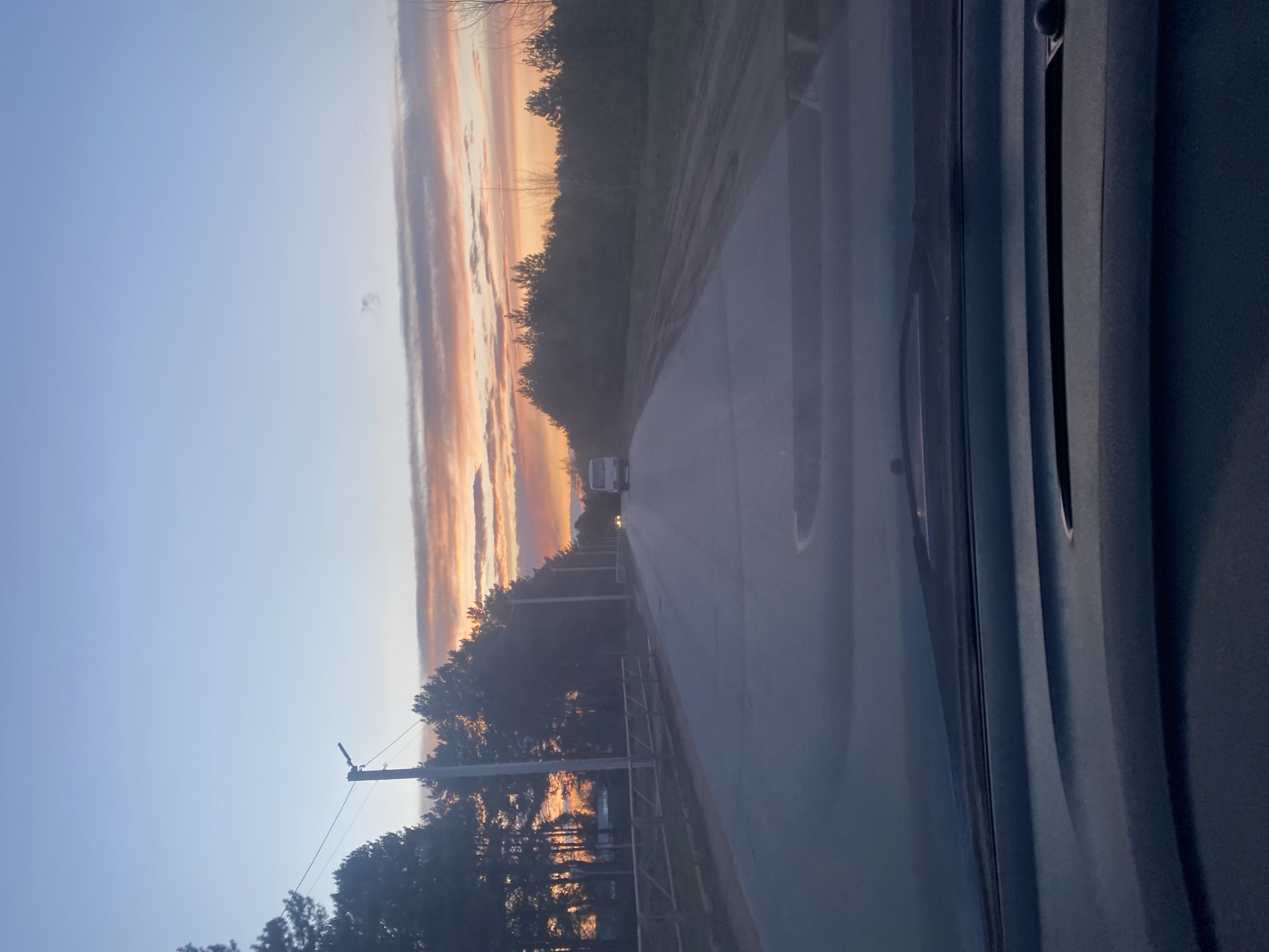 This image captures a tranquil road scene at twilight, viewed from inside a vehicle in Chaikovsky, Russia. The perspective is from a side window, with a dark, textured car interior panel visible on the right. The sky is a prominent feature, displaying a beautiful gradient from a soft blue at the top to warm, fiery oranges, pinks, and purples along the horizon, where long, streaky clouds are illuminated by the setting or rising sun.
A paved road occupies the central and lower portions of the frame, extending into the distance. On the left side of the road, a dense line of dark trees, possibly evergreens, forms a natural boundary, with utility poles and wires interspersed among them. Further along this side, a fence or guardrail is visible. On the right side of the road, the landscape appears less dense with foliage.
In the far distance on the road, a white vehicle, likely a truck or van, is visible, with its headlights illuminated, suggesting the low light conditions of either sunrise or sunset. The overall atmosphere is peaceful and calm, highlighted by the soft, warm light. No people are explicitly visible, and there is no discernible text within the image.