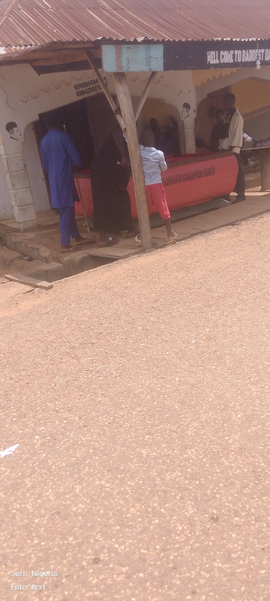 A low-angle, exterior medium shot shows a wooden structure with a corrugated metal roof, possibly a roadside stall or shop. In the background, a red object, possibly a table or display, has white text that reads "STANDARD CHAMPION BORD". Above the structure, a sign reads "HELL COME TO BADDEST BA". Several people are present: a man in a blue kaftan stands to the left, a person in black attire stands in the center, a boy in a light blue shirt and red shorts stands to the right, and another person in a light-colored shirt and dark pants stands further to the right, behind the boy. The ground in the foreground is a dirt or gravel path. The overall scene suggests a commercial area in an urban or semi-urban environment.