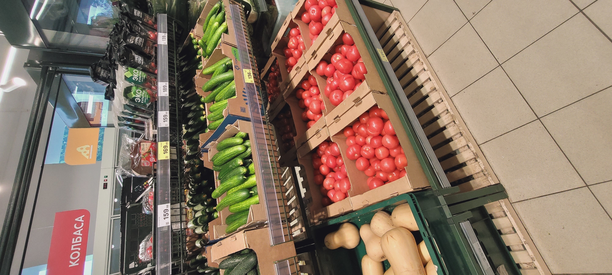 The image shows the produce section of a grocery store. On the left, there is a refrigerated display case with sausage and other products. Next to it is a display of cucumbers arranged in cardboard boxes. The price labels are visible, showing prices like 159.99, 169.99, 189.99, 139, and 136.19. Next to the cucumbers, there are cardboard boxes filled with tomatoes. In the bottom right corner, there is a green crate with squash. The floor is tiled with beige square tiles. The location is in a city of Unknown, Unknown.