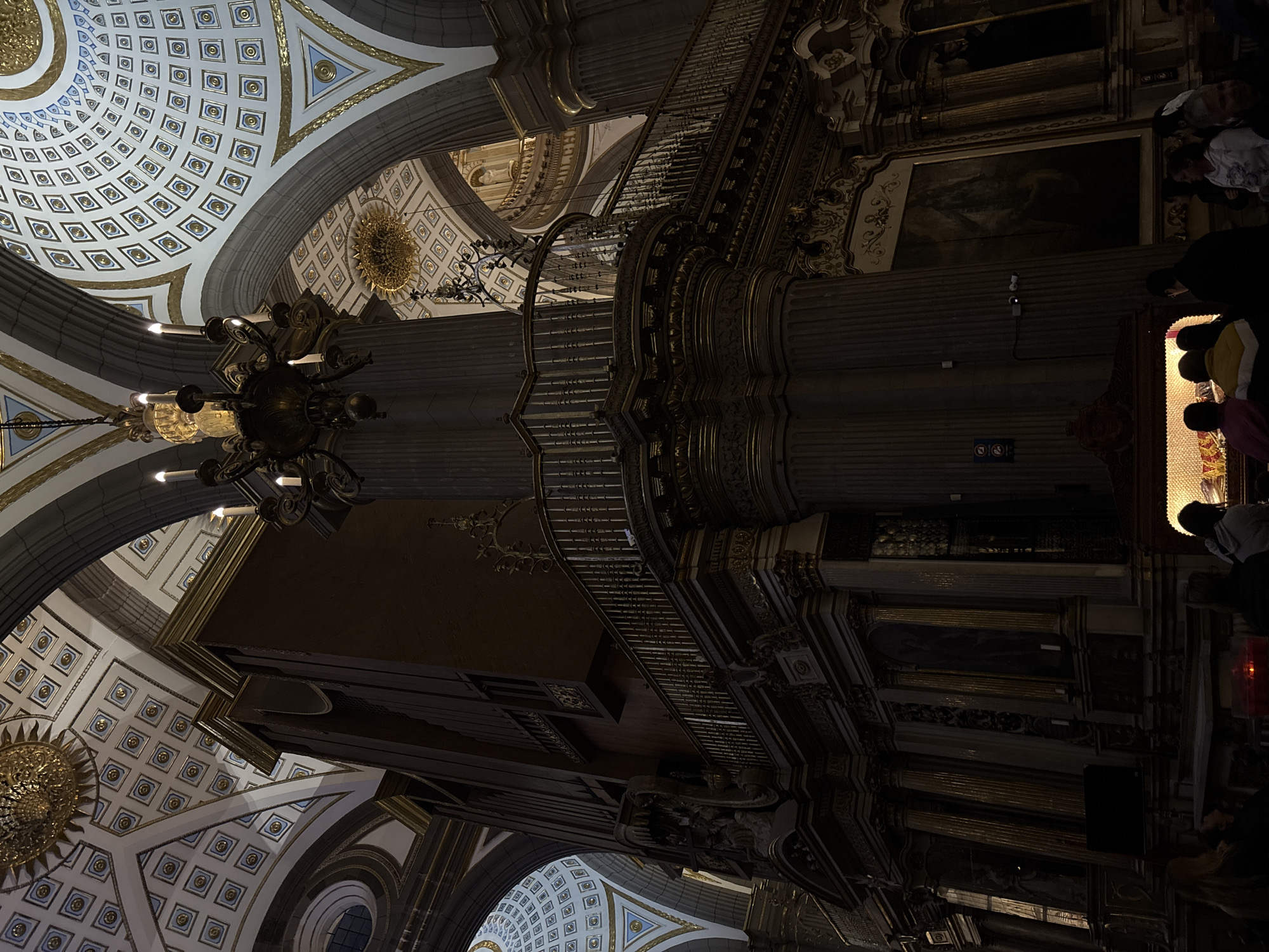 The image shows the interior of Puebla Cathedral in Puebla, Mexico. The view is angled upwards, showcasing the ornate ceiling with geometric patterns and golden accents. A large, multi-tiered chandelier hangs in the foreground. The architecture includes massive columns and an elaborate organ structure, both adorned with intricate carvings and details. On the right side of the image, there are people present, possibly viewing a religious display or monument. The lighting is dim, emphasizing the grandeur and historic nature of the cathedral.