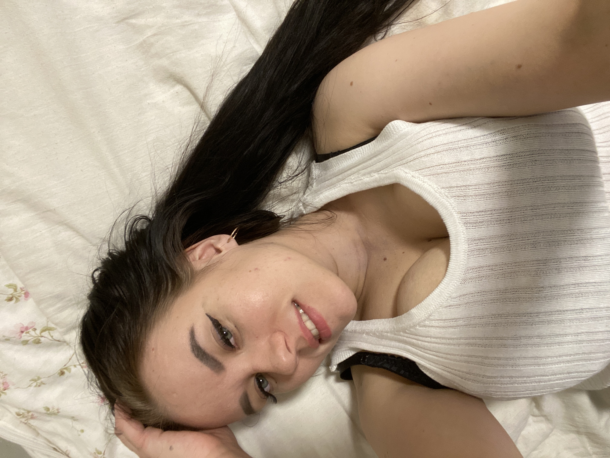 A woman with long, dark hair and fair skin is lying on a bed with a white and pink floral patterned sheet. She is wearing a white ribbed tank top with a low neckline. She is smiling and looking at the camera. Her head is resting on her left hand, which is visible on the bed. The background is plain and uncluttered. The photo does not provide information to determine if this image was taken in Chaikovsky, Russia.