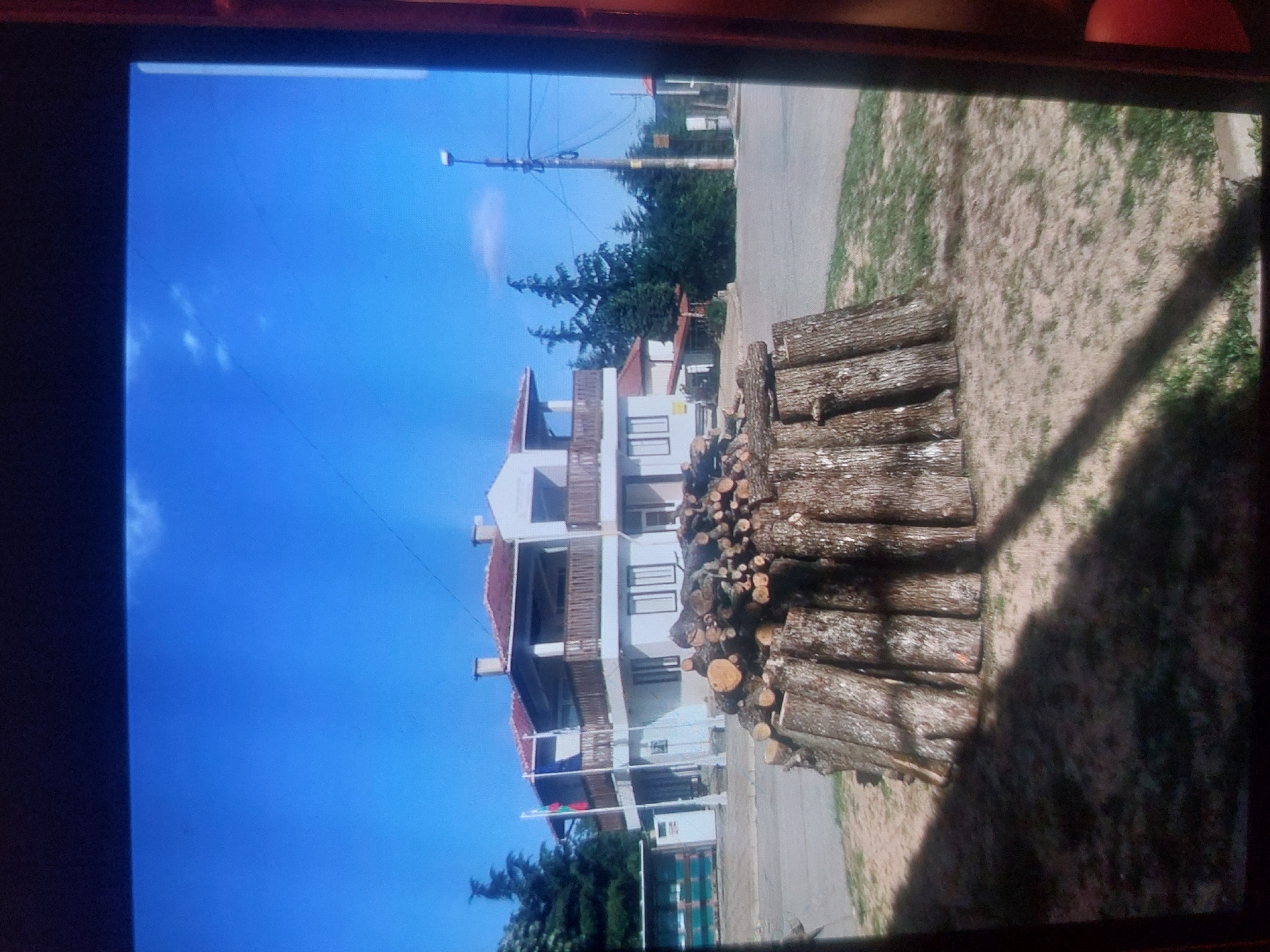 The image depicts a stack of logs next to a paved road, likely for firewood, in an outdoor environment under a clear blue sky with some light clouds. In the background, there is a three-story white building with balconies, possibly a hotel or apartment complex, as indicated by the presence of several windows and railings. A street lamp and power lines are also visible. Further in the background, trees and foliage contribute to the setting, suggesting a suburban or semi-rural location within Tsarevo, Bulgaria. A Bulgarian flag can be seen at the base of the building.