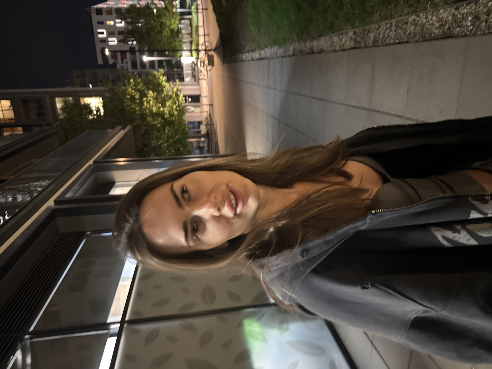 A medium shot shows a woman with light brown hair, standing outside at night. She is wearing a dark-colored jacket over a light-colored top. The background is an urban street scene with buildings, street lights, and trees. The woman is the primary subject, and the urban background provides context to the scene. The location is Unknown, Unknown.