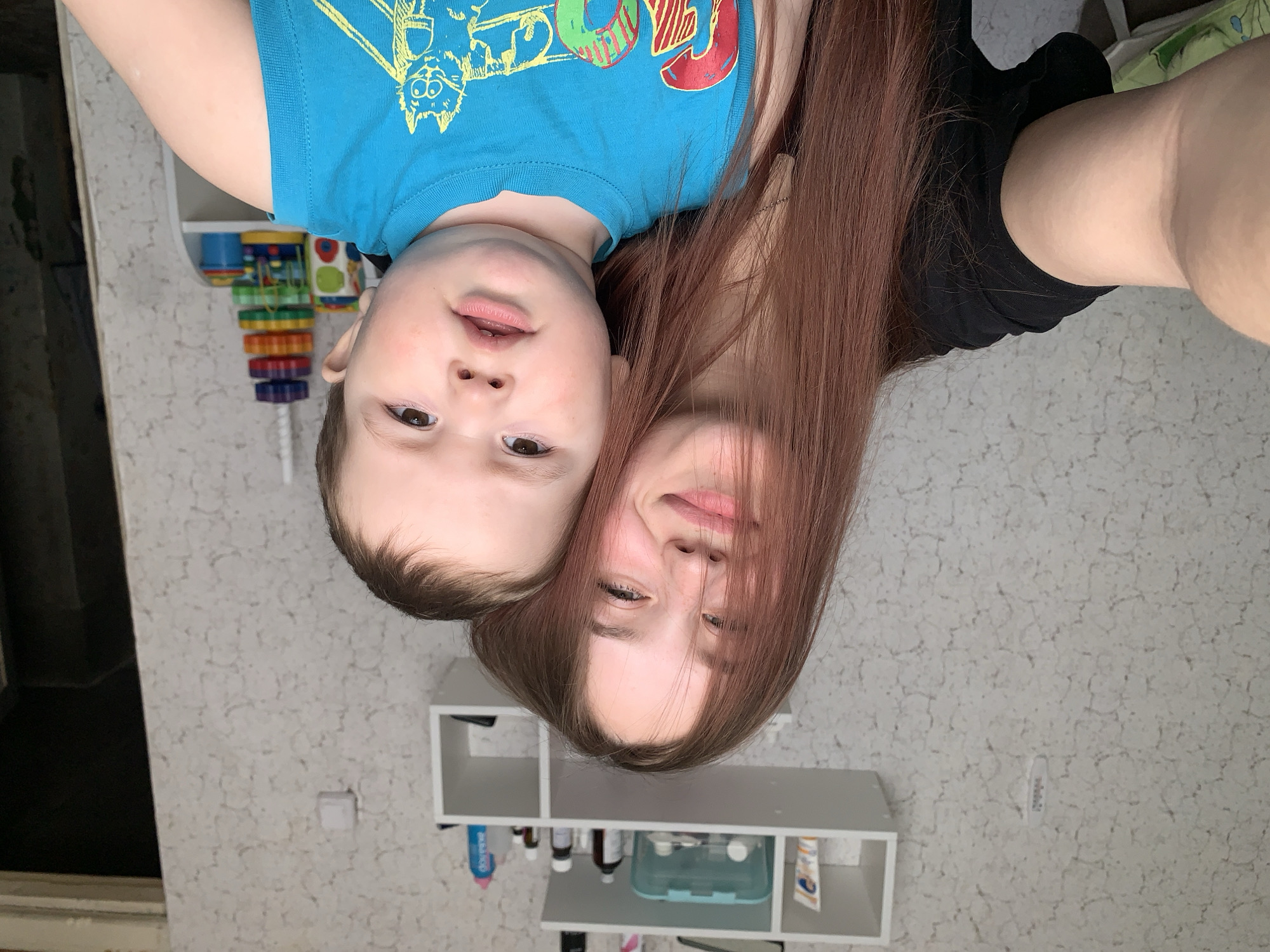 Two individuals, a young child and a young woman, are positioned close together in an indoor setting. The child is wearing a blue t-shirt with cartoon graphics. The woman has long brown hair. They appear to be taking a selfie. The background includes a textured wall, a white shelf with various bottles and containers, and a children's toy hanging on the wall. Based on the location mentioned, the image was likely taken in the city of Фоки, Russia.