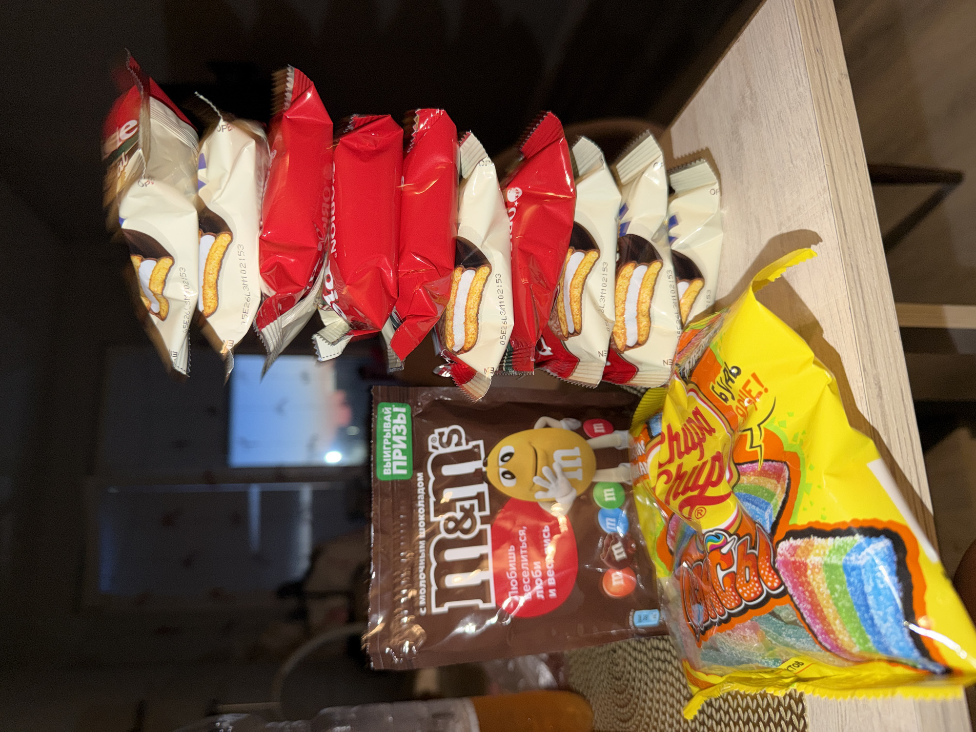 The image shows a collection of snack items arranged on a wooden surface, likely a shelf or countertop, in a domestic setting. The items are predominantly sweet treats.
In the foreground, a brown packet of M&M's is visible, featuring the brand's iconic characters and text in Russian, including "ВЫИГРЫВАЙ ПРИЗЫ!" (Win prizes!) and "с молочным шоколадом" (with milk chocolate). Beside it, a brightly colored yellow bag of Chupa Chups sour straws is prominently displayed, showcasing a rainbow design and the brand's logo.
Behind these, a series of individually wrapped snacks are hanging or attached to a surface. These appear to be chocolate biscuit snacks, with some wrappers being red and others a cream color with images of the biscuits. The wrappers have visible printed text, some of which is legible due to the provided crops, indicating batch numbers and possibly expiry dates.
The background is blurred, but suggests an indoor environment, possibly a kitchen or pantry, with some reflective surfaces and dimly lit areas. The lighting appears to be artificial, suggesting the photo was taken indoors, potentially at any time of day. The overall impression is of a casual arrangement of snacks, perhaps prepared for a snack break or gathering.