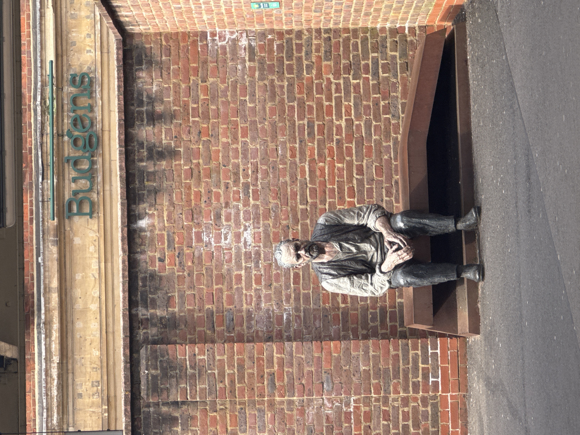 A weathered, life-sized statue of an elderly, bearded man is seated on a dark, angular metal bench in front of a red-brown brick wall. This scene is situated at Station Approach, Woking, United Kingdom, beneath a light-colored stone fascia displaying the green 'Budgens' retail sign. The statue, depicting a man in a shirt, waistcoat, and trousers with clasped hands, gazes forward with a somber expression. The brickwork shows areas of dark staining and light efflorescence. A small vertical blue-green sign with white numbering '3 11' is visible on the adjacent right wall section. The foreground consists of dark asphalt pavement.