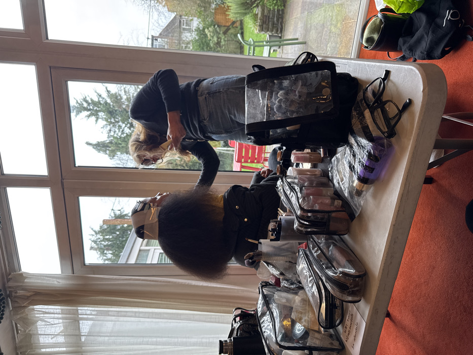 The image depicts an indoor scene within a residential property in London, United Kingdom, overlooking a garden. In the foreground, a white table is covered with numerous clear plastic cosmetic bags containing various makeup products, alongside a cylindrical brush holder filled with brushes, a purple bottle, and a pair of black reading glasses. A dark thermos is also visible on the table's left.
To the left, a person with dark, voluminous hair is seated, facing away from the camera in profile. Their hair at the crown is secured with what appear to be rollers or paper. They are wearing a dark jacket, possibly over a yellow garment. A second individual, with long, light-colored hair and glasses, stands to the right, wearing a dark long-sleeved top and blue jeans. This individual is leaning in, holding a light-colored tool, and actively applying makeup or styling to the seated person's face.
The background consists of large, white-framed glass doors and windows, revealing an outdoor garden scene under an overcast sky. Visible outside are green grass, several trees (some bare), red outdoor chairs, and a distant house. The indoor flooring is covered with orange carpet.