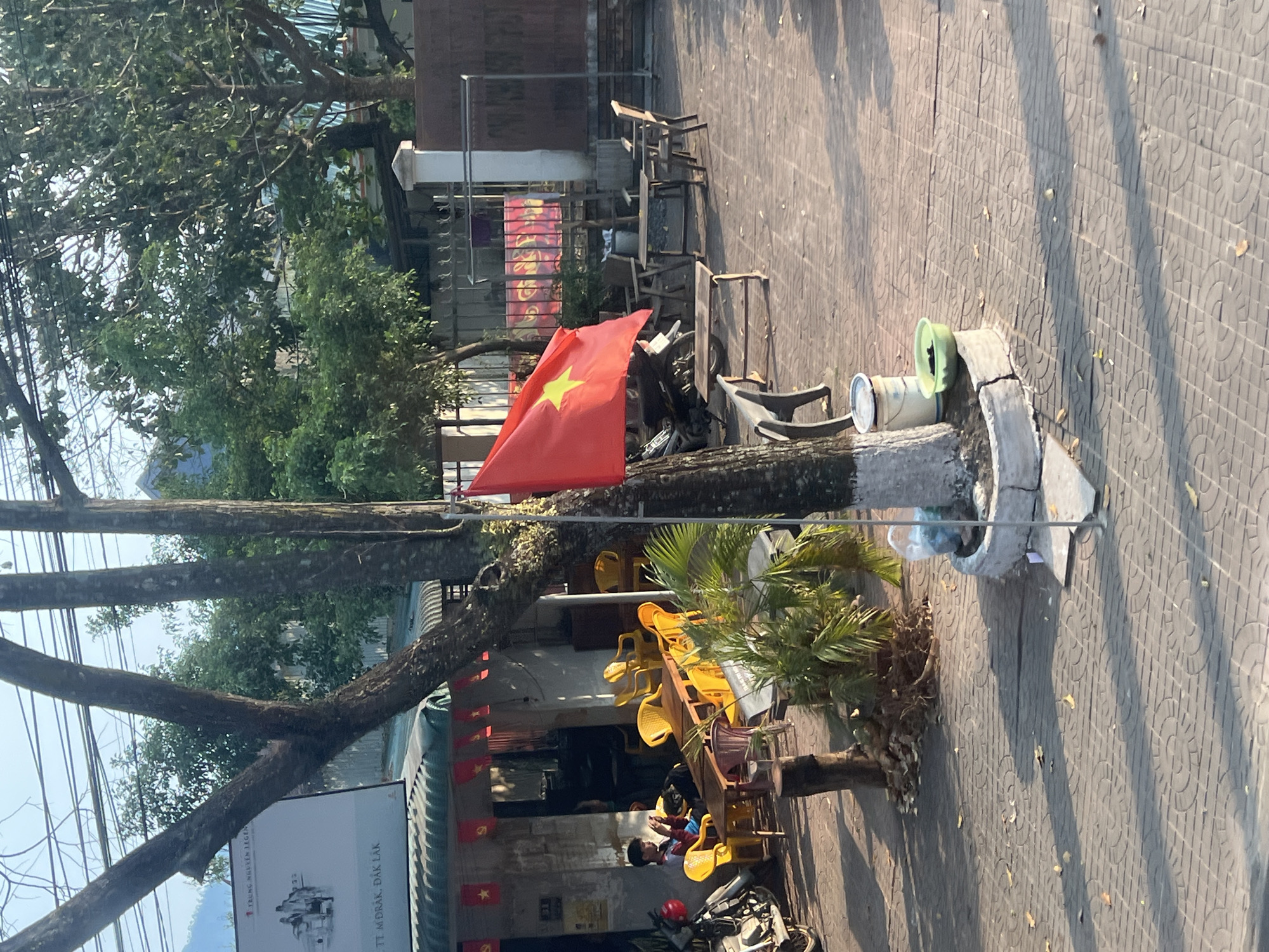 A street scene in M'Drắk, Đắk Lắk, Vietnam. A Vietnamese flag is displayed on a tree. Yellow chairs are stacked outside a building adorned with more Vietnamese flags. A child sits among the chairs. A parked motorbike is partially visible. A sign for "Trung Nguyên Legend" is also visible. The ground is paved with patterned tiles.