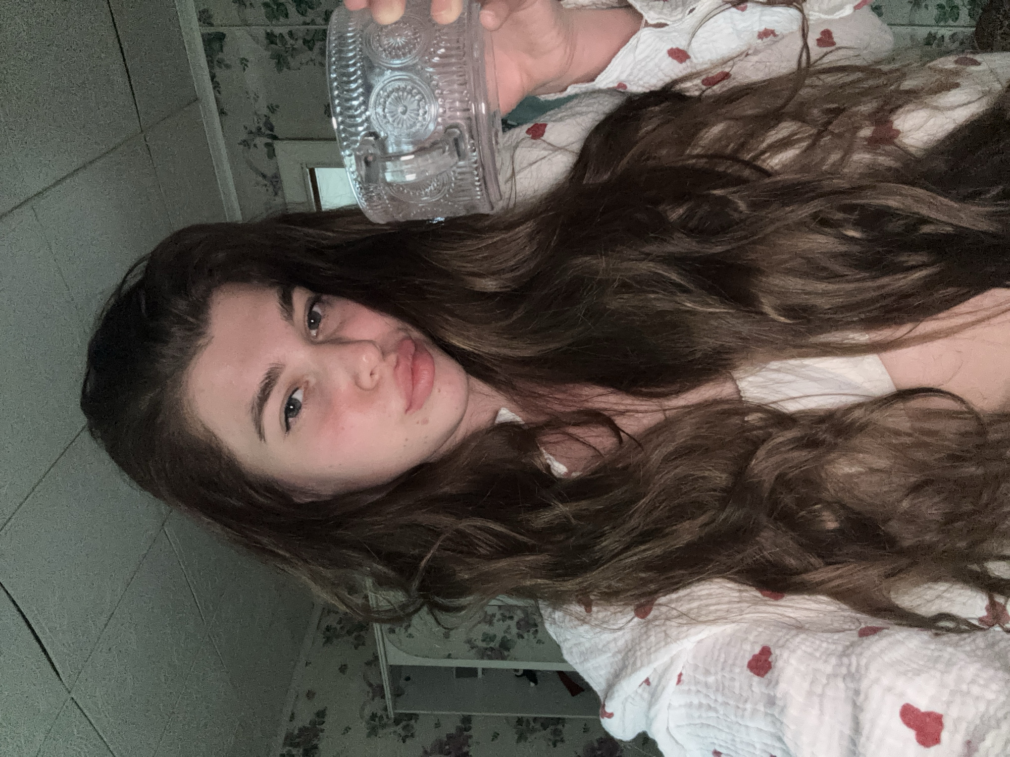 A young woman with long brown hair is lying down, holding a clear, ornate glass container above her head. She is wearing a white top patterned with red hearts. The background includes a tiled wall and patterned wallpaper. The location appears to be indoors, possibly a bedroom. There is no clear indication that the location is the city of Мещёры, Russia.