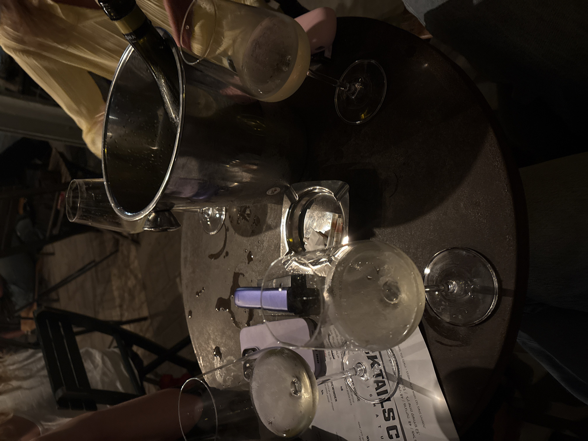 In the evening, at an outdoor establishment in Tel-Aviv, Israel, a round, dark-colored table is the central focus. Several glasses of white wine, some partially filled, are scattered across the table. A metal ice bucket with a bottle of wine inside stands prominently. An ashtray, containing cigarette butts, rests near one of the wine glasses. A cell phone is also visible on the table, next to a small, purple electronic device. In the background, blurred figures of people are seated at chairs, some facing away from the camera. The overall ambiance suggests a casual social gathering.