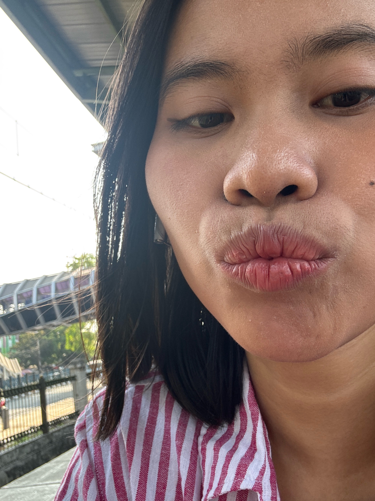 A woman with dark hair and fair skin is posing for a close-up photograph, puckering her lips. She is wearing a pink and white striped shirt. In the background, out of focus, is a modern elevated pedestrian walkway with a purple railing, typical of infrastructure found in the city of East Jakarta, Indonesia. The sunlight suggests it is daytime.