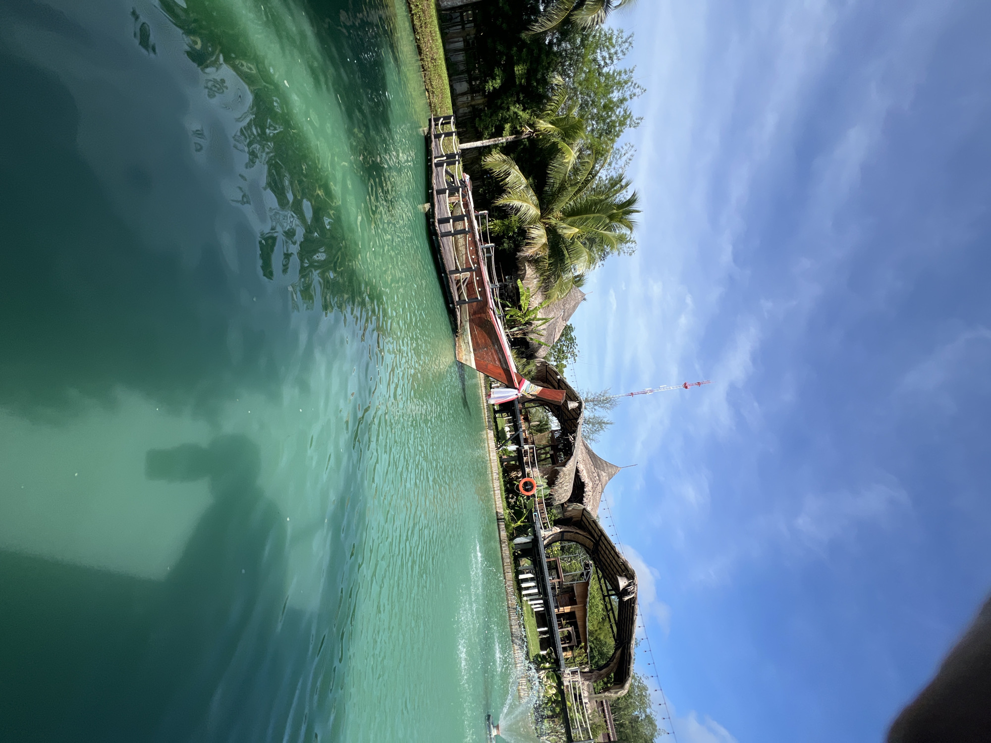 This media file depicts a tranquil, tropical waterside scene in Tambon Pa Kae Bo Hin, Thailand. The primary subjects include a wooden boat moored to a dock, several thatched-roof structures, lush greenery with palm trees, and a bright blue sky with scattered clouds. The water is a clear, vibrant turquoise, reflecting the surrounding environment.
The boat appears to be a traditional longtail boat, with its distinctive wooden hull and decorative elements. It is positioned alongside a rustic wooden pier that extends into the water. Behind the pier, the thatched-roof buildings suggest a resort or restaurant, with open-air designs characteristic of tropical architecture. The presence of a life preserver ring attached to a railing on one of the structures implies a focus on safety and water activities.
The scene is bathed in bright sunlight, indicating it is daytime, likely mid-morning or afternoon, given the clear sky and the quality of light. The weather appears to be warm and pleasant, typical of a tropical climate.
Notable details include the reflection of a person in the water, possibly the photographer, which adds a personal element to the otherwise scenic view. The overall emotion conveyed is one of peace and relaxation, inviting viewers to imagine themselves in this idyllic setting. The location cues, such as the specific type of vegetation and architecture, strongly suggest a Southeast Asian tropical environment, specifically Thailand as indicated by the provided location context. No visible text is present in the image.