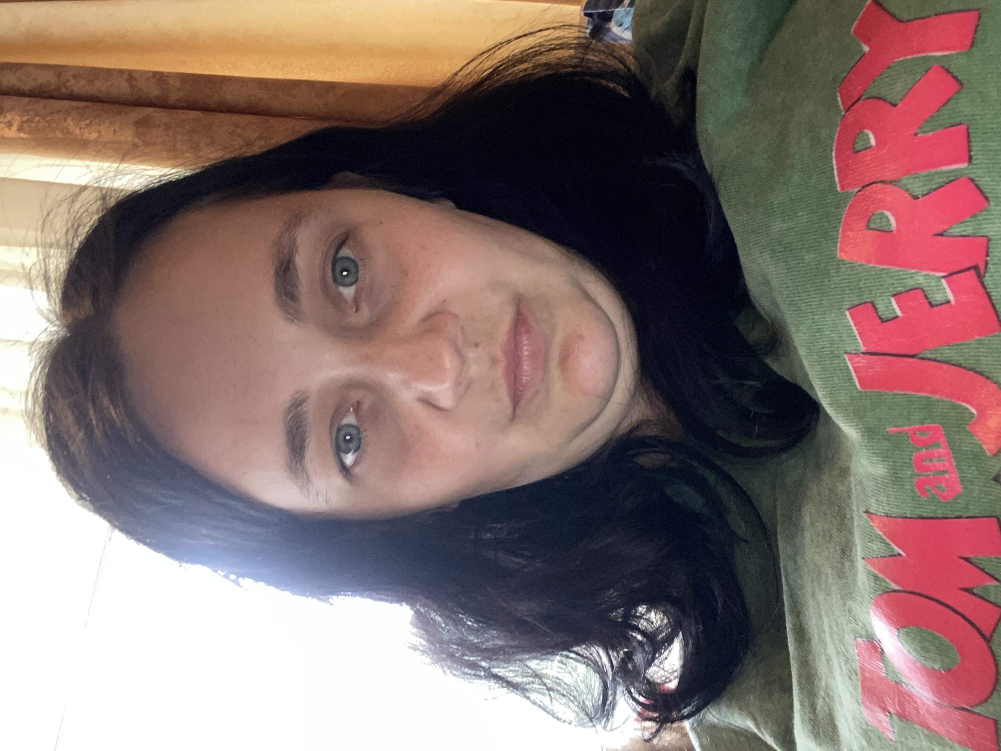 A medium shot captures a woman with dark, wavy hair and blue eyes looking directly at the camera. She has fair skin and appears to be wearing a green t-shirt with large red lettering that reads "TOM and JERRY". The background features a bright, diffused light source on the left and orange curtains on the right. The lighting suggests the photo may have been taken indoors, possibly near a window.