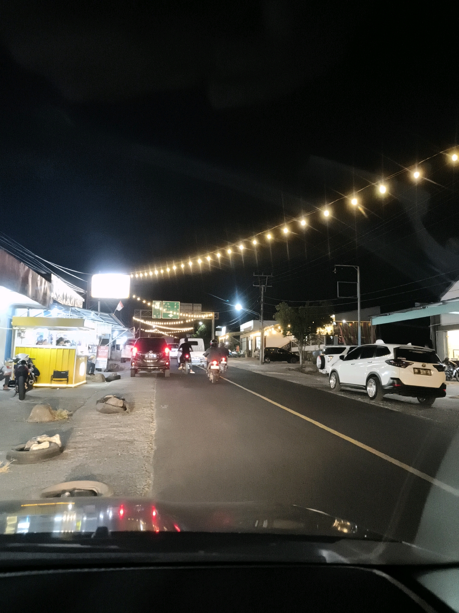The image depicts a street scene at night in what is identified as Talikuran, Indonesia. A string of yellow lights illuminates the upper portion of the frame, creating a festive or decorative atmosphere. The street is lined with various establishments, including a yellow food stall with customers, and other buildings with illuminated signs and storefronts. Several vehicles are present, including cars, motorcycles, and what appears to be a small truck. Two individuals are seen on motorcycles in the middle of the road, and at least one person is visible at the food stall. Tires and rocks are placed along the side of the road. The overall impression is that of a lively commercial area at night.