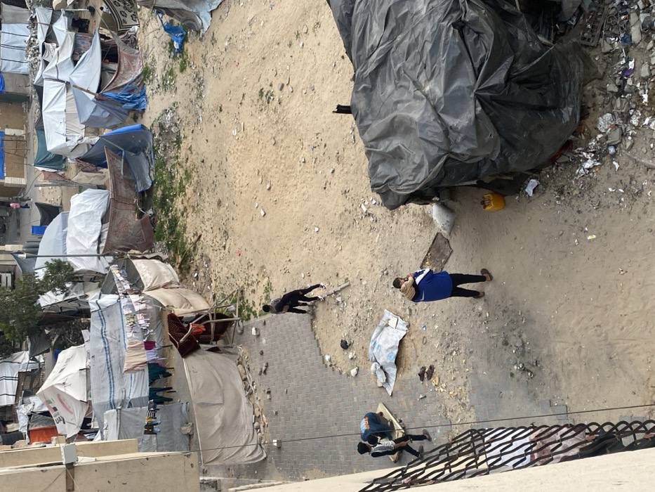 The image captures an overhead view of an informal settlement or refugee camp, looking down from an elevated position, possibly a building or balcony, as indicated by a black railing visible in the lower right foreground. The scene is dominated by makeshift structures and a dusty, somewhat barren landscape.
The ground consists primarily of light-brown dirt and sand, interspersed with patches of sparse, dry green vegetation. Sections of this ground are littered with debris, including small white fragments, plastic, and general waste. A paved area, appearing to be made of cobblestones, is visible in the lower-middle portion of the image.
On the left side of the frame, numerous temporary shelters or tents are densely packed together, extending towards the background. These structures are constructed from a variety of materials, predominantly white and blue tarpaulins, as well as patterned fabrics that resemble carpets or blankets. Clothes are seen hanging on lines strung between some of these shelters, drying in the open air.
In the upper-right quadrant, a large, dark, possibly black or dark blue, tarp-covered mound or structure is prominent. Its crinkled texture suggests a substantial piece of fabric covering an object or area. Near the base of this mound, on the dirt ground, a small, bright yellow plastic container (possibly a jerry can) is visible.
Several individuals are present in the lower half of the image:
* Towards the center-right, a person wearing a blue top and dark trousers stands on the dirt ground, holding a small child or infant wrapped in a light-colored blanket or clothing.
* To their left, in the mid-left section, another individual in dark clothing (black top and pants) is standing, leaning slightly forward.
* In the lower-right foreground, near the railing, two more people are visible on the paved ground. One is wearing a striped top and dark pants, while the other is partially obscured but also appears to be in dark attire.
The scene depicts daily life in a challenging environment. The natural lighting suggests it is daytime, though the light is diffused, possibly indicating an overcast sky or a time of day with less direct sunlight. Faint, unreadable text, possibly "UAE" or "AID," can be seen on a blue stripe on one of the white tent fabrics in the lower-left cluster.