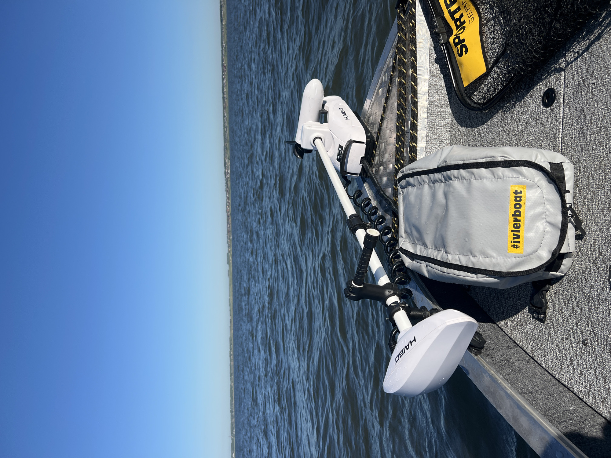 A perspective shot from the front of a boat on a body of water under a clear blue sky. The boat's bow, made of gray textured material, is visible in the foreground, with a metal railing along the edge. Mounted on the boat is a white electric trolling motor with the brand name "HAIBO" visible on its housing. Next to the motor is a gray bag with a yellow tag that reads "#ivlerboat". To the right of the bag is a black fishing net with a yellow handle labeled "SPORTEX GERMANY". In the background, a calm body of water stretches to the horizon, where a distant shoreline with trees and houses can be seen. The location is the city of Novopichugovo, Russia.