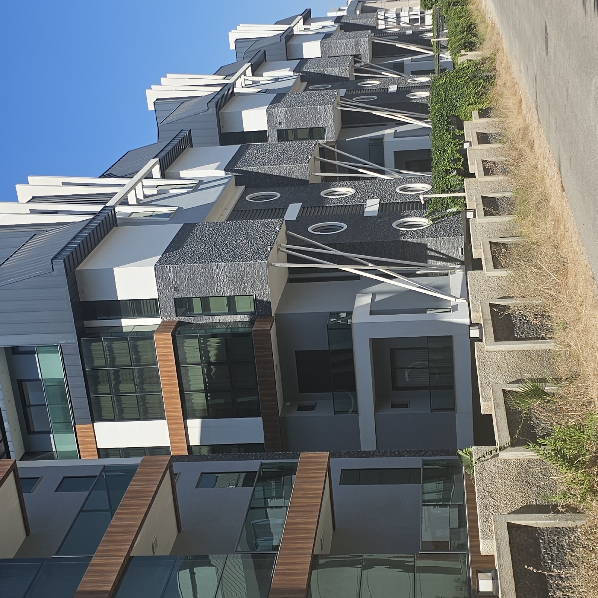 The image shows a row of modern residential buildings in Alanya, Turkey, with distinctive architecture featuring angled dark gray roofs, white and dark gray facades, and large glass windows. The buildings are multi-story with balconies and decorative elements such as circular windows and angled support beams. In the foreground, there is a low concrete fence with rectangular openings filled with rocks, and dry vegetation. A green iguana is visible on top of the fence on the left side of the image. The buildings appear to be located in a developing area, possibly a residential complex, with a paved road to the right. The sky is clear and blue.