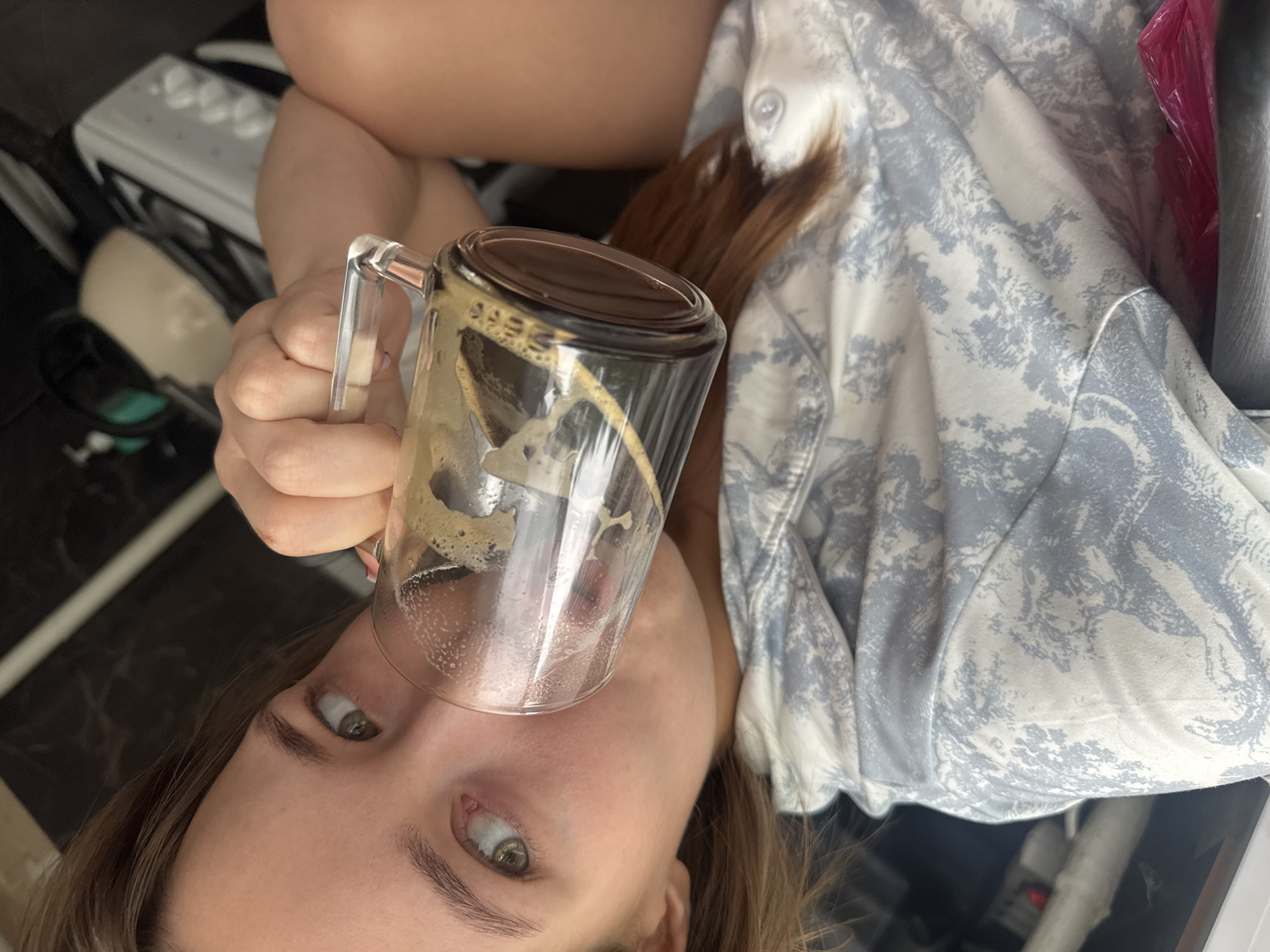 A close-up shot shows a young woman with light brown hair and green eyes drinking from a glass mug. She is wearing a light blue and white patterned shirt. The mug contains a dark liquid, possibly coffee. The background is out of focus, but appears to be an indoor setting with some kitchen appliances visible. The image was taken in Chaikovsky, Russia.