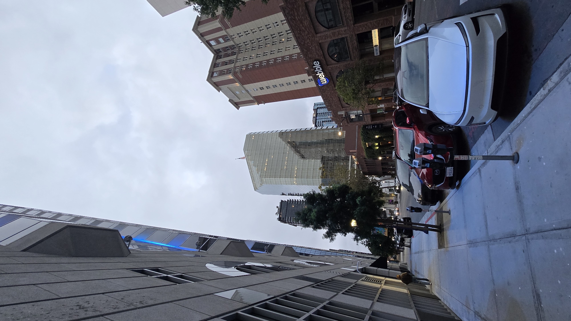This is an eye-level, wide-angle, outdoor shot of a street scene in downtown San Diego, California, USA, under an overcast sky. The perspective is looking up and down a city street lined with multi-story buildings. On the left, a modern building with a grey facade and large windows dominates the foreground. To the right of that building is a sidewalk with people walking and several parked cars, including a white electric vehicle in the immediate foreground, a red SUV, and a dark sedan. Across the street, there are several other buildings, including a brick building with a "webble" logo and a tall, glass-fronted skyscraper with an American flag on top. The street appears to be well-lit with streetlights and lights from the buildings. The overall impression is of a bustling urban environment.