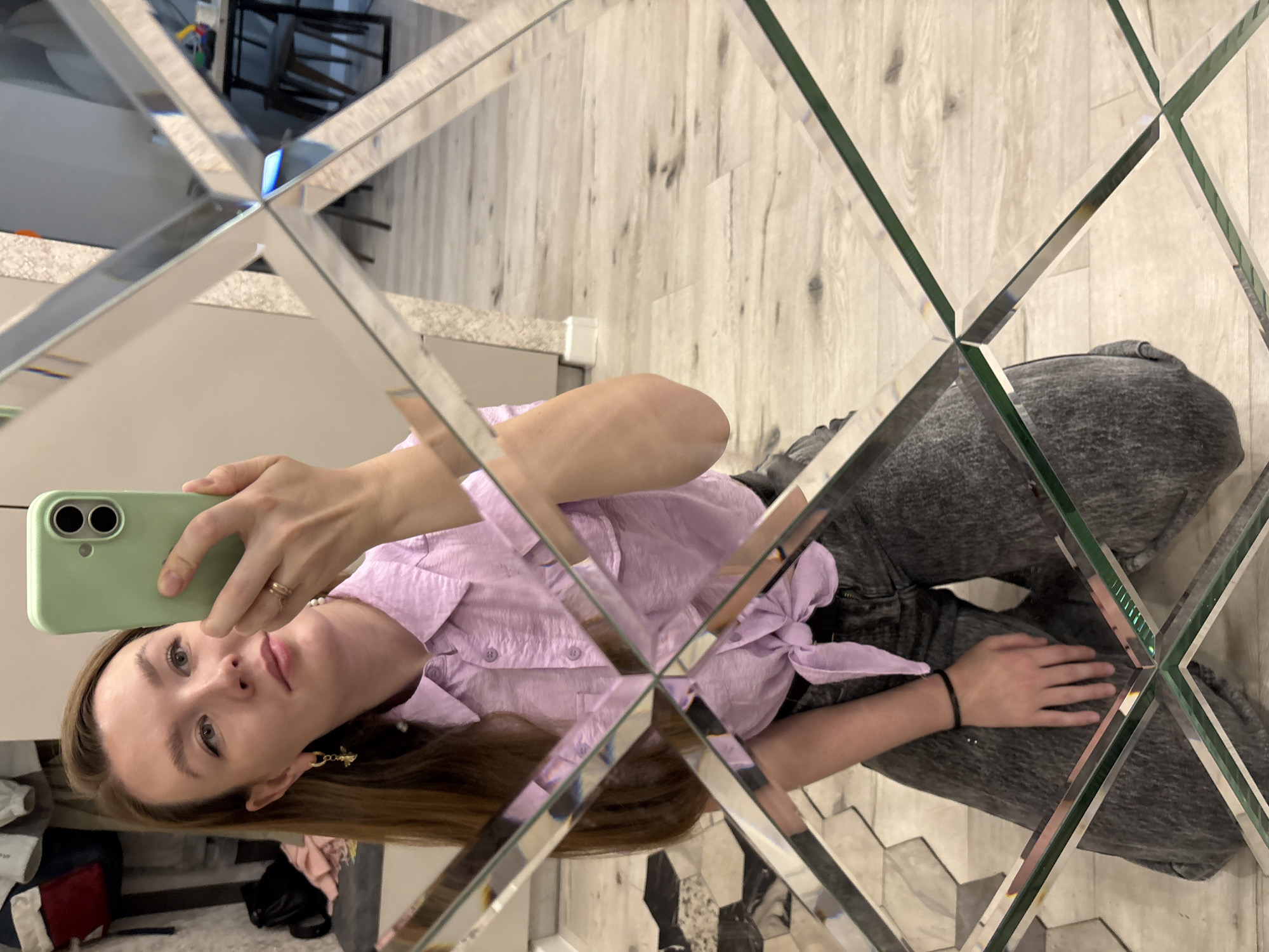 A young woman is captured in a mirror selfie, seen through a large decorative mirror composed of beveled, diamond-shaped segments. The image is oriented sideways, with the woman's head to the left. She has long brown hair and is wearing a light purple short-sleeved blouse with a tie detail at the waist, paired with dark grey sweatpants.
She holds a light green smartphone with two camera lenses, pointing it towards herself to take the photo in the mirror. A gold ring is visible on her right hand, and a gold earring (possibly shaped like a bee or insect) is on her left ear. She also wears a pearl necklace. Her expression is neutral as she looks directly at the camera.
The background, visible through and around the mirror segments, features a light-colored wooden floor and a light-toned countertop. Reflections in the mirror distort parts of the room, showing what appears to be dark furniture or a chair in the upper left segment. The setting suggests an indoor environment, likely a modern living space or hallway, illuminated by natural light during the daytime. There is no visible text in the image.