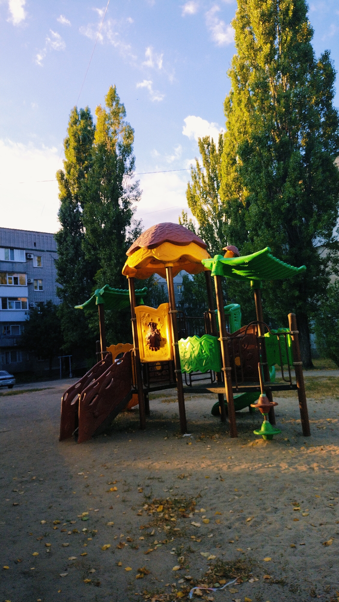 The image depicts a children's playground in Horishni Plavni, Ukraine, bathed in the warm light of either sunrise or sunset. The playground features a multi-colored structure with brown slides, yellow and orange roof elements, and green plastic decorative pieces. A small, dark object, possibly a toy animal, is visible on a yellow panel within the playground. In the background, a grey apartment building with multiple windows stands against a backdrop of tall, slender trees with green foliage. The ground of the playground is covered in sand and scattered fallen leaves, suggesting an autumn setting. The sky above is blue with scattered white clouds and a thin, diagonal line, possibly a contrail or a string, stretches across it. A portion of a grey car is visible on the left side of the image.