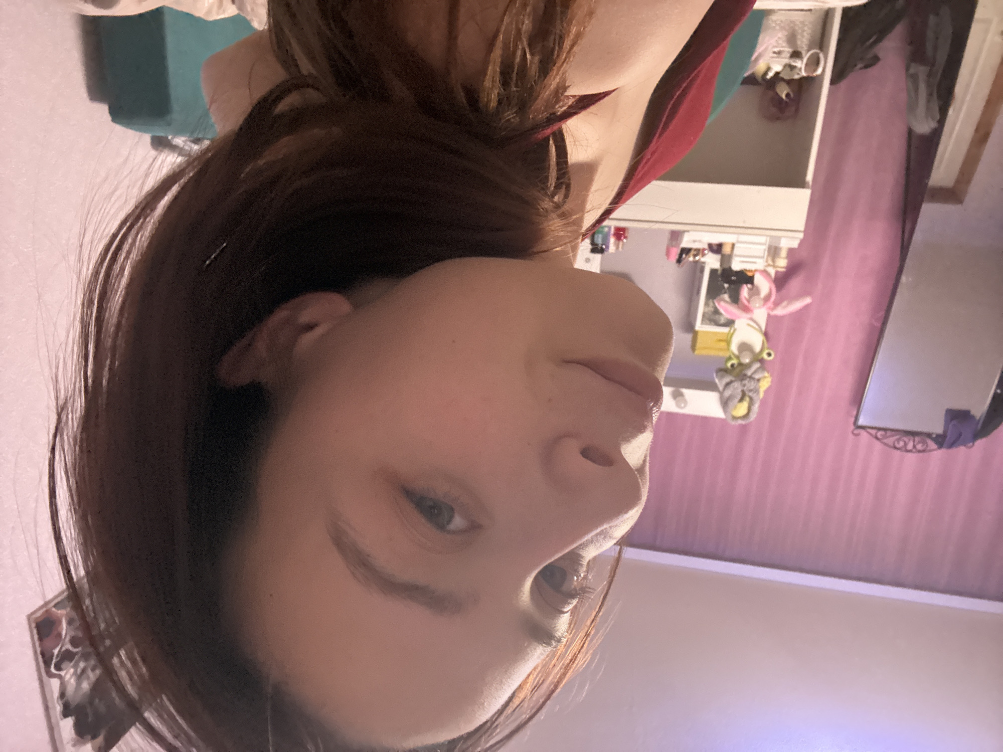 The image displays a close-up selfie of a young person, captured indoors from a low, tilted angle. The individual has long, dark brown hair that frames their face, and light skin. Their right eye is open and directed towards the camera, while their left eye is mostly obscured by hair. They wear a reddish-maroon top, with part of their shoulder and upper chest visible. Their expression is neutral to contemplative.
The background reveals a personal room, likely a bedroom. Behind the person, a light-colored dresser or shelving unit holds various objects, including a distinctive plush toy resembling a frog with a pink bow. To the right, a mirror with a decorative, possibly wrought-iron, frame reflects parts of the room and the person's hair. The wall is adorned with light purple or pink wallpaper, which appears to have a textured or subtle striped pattern, separated by a white trim from a lighter ceiling or upper wall section. A teal object is partially visible in the distant background. The lighting suggests artificial indoor illumination. No text is visible. The context for this image is Прикамский, Russia.