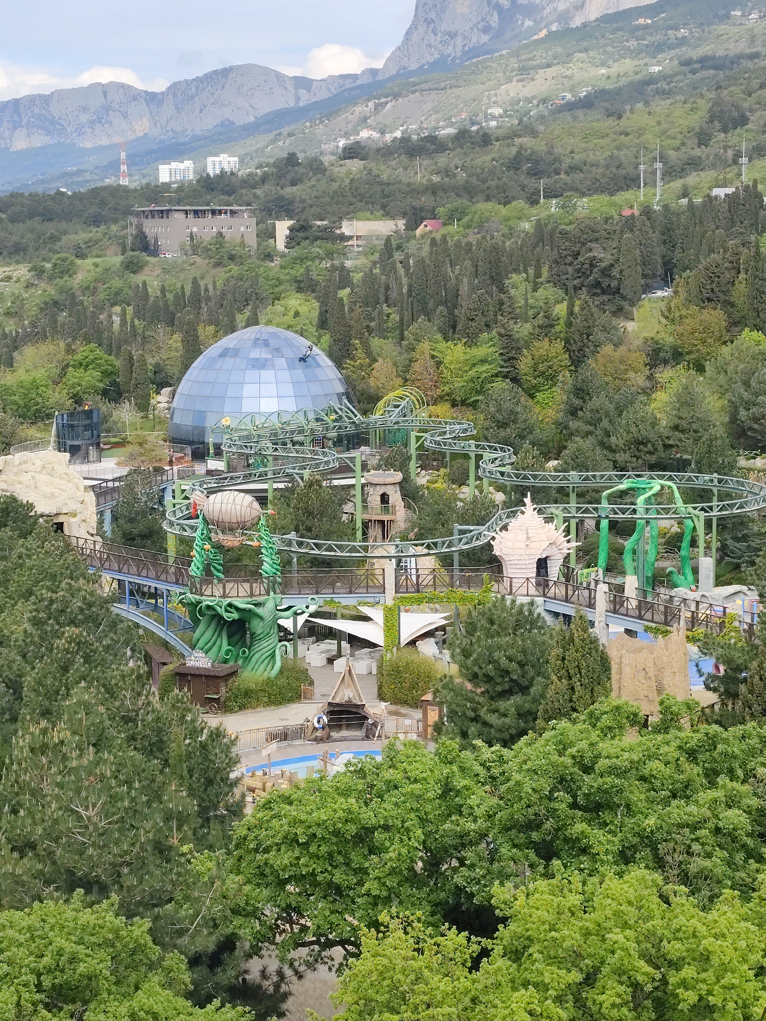The image depicts a high-angle view of an amusement park in the city of Симеизский поселковый совет, Ukraine, set against a backdrop of lush greenery and mountainous terrain. The central focus is a complex of attractions, including a domed building constructed of glass, a winding roller coaster with green tracks, and various thematic elements such as fantasy-style buildings, artificial rock formations, and faux vegetation, specifically, oversized beanstalk structures. Below the park infrastructure, one sees a swimming pool and other structures. The area is surrounded by dense trees. In the background, beyond the park, is a cluster of buildings and distant mountains under a cloudy sky.