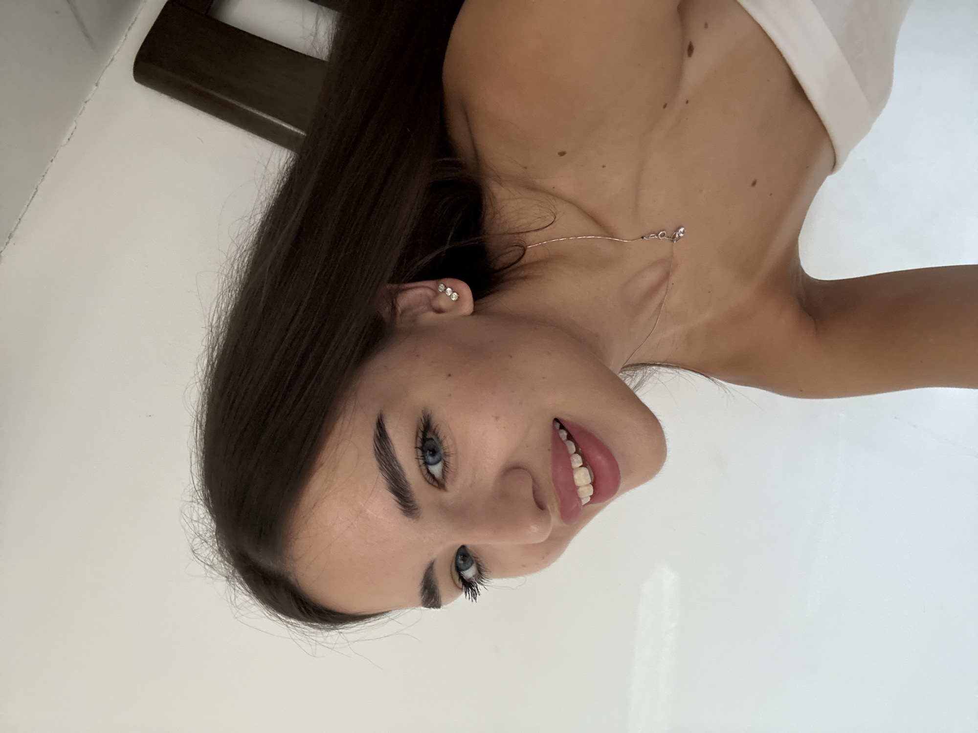 A young woman with long, dark hair and blue eyes is shown in a close-up, angled portrait. She is smiling with her mouth open, revealing her teeth. Her head is tilted to her left, and her gaze is directed slightly upwards. She is wearing a simple white garment off one shoulder, a delicate necklace with a small pendant, and a cluster of three small earrings in her right ear. The background is a plain white wall, with a portion of a dark brown wooden object, possibly furniture, visible on the right side of the frame. No specific location in Chișinău, Moldova, can be identified from the image.