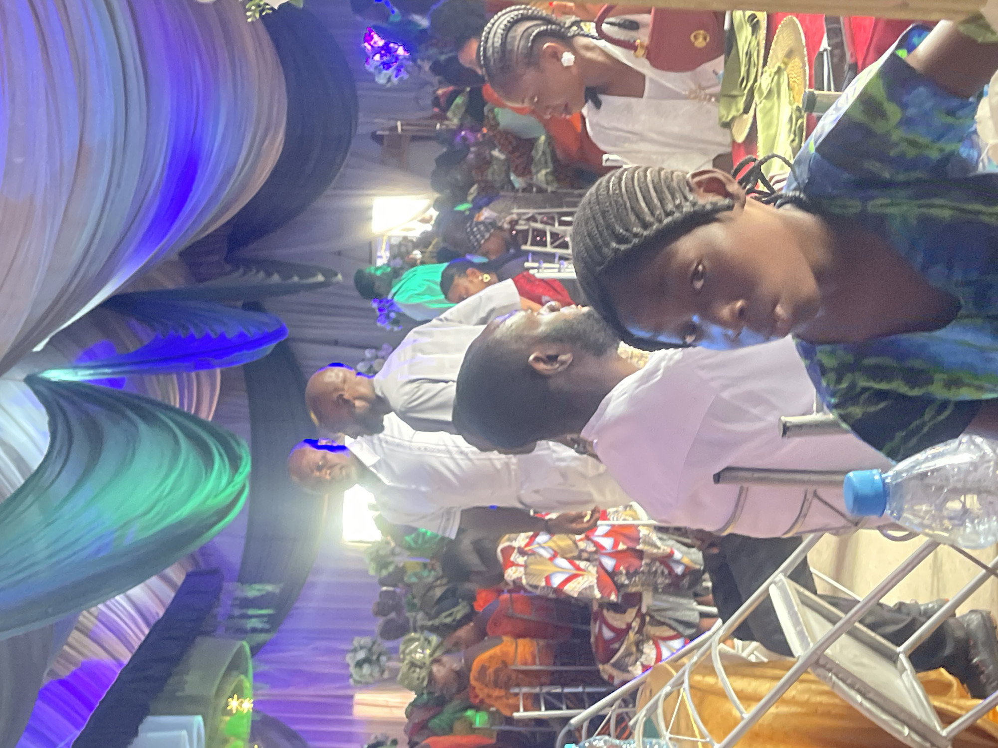 A densely packed indoor event is in progress in Jos, Nigeria. The event appears to be a formal gathering or celebration, as evidenced by the decor and attire of the attendees. The space is decorated with draped fabrics, including blue, green, and beige hues, indicating a coordinated color scheme. Attendees wear formal attire, with several individuals dressed in white garments. One person stands out with a vibrant blue and green patterned shirt. There is a water bottle on the metal chair. Some attendees wear traditional head coverings. The overall atmosphere suggests a festive and celebratory occasion.