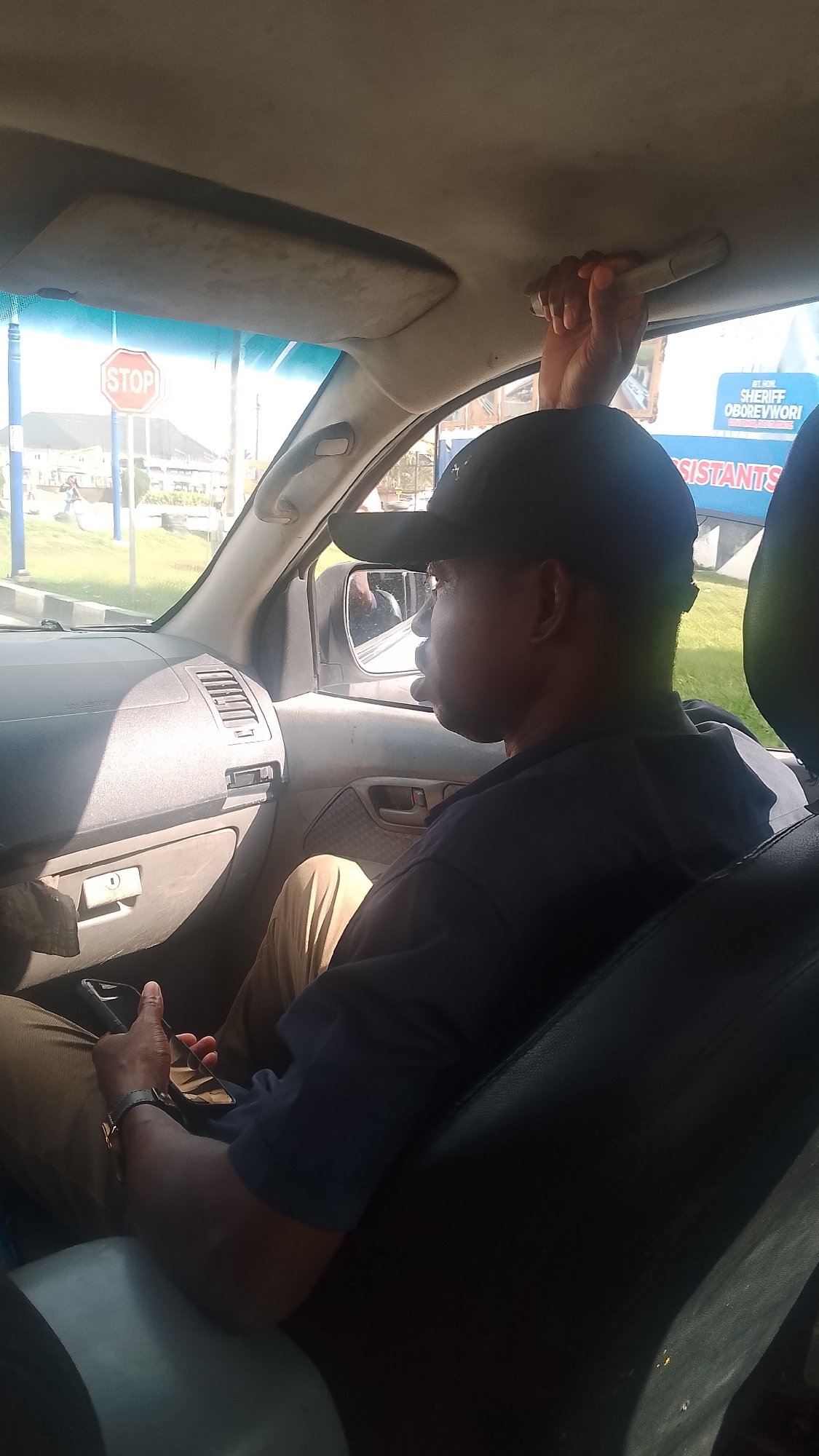 A man in a black t-shirt and cap sits in the passenger seat of a car. He is holding a phone in his left hand, and his right hand grips the car's handle above the window. Outside the car window, a "STOP" sign is visible on a blue and white pole. A building is in the background, and a sign with "RT. HON. SHERIFF OBOREVWORI" is also visible. The location is Orerokpe, Nigeria, based on the sign mentioning "OBOREVWORI". The car's interior, including the dashboard, seats, and rearview mirror, is visible.