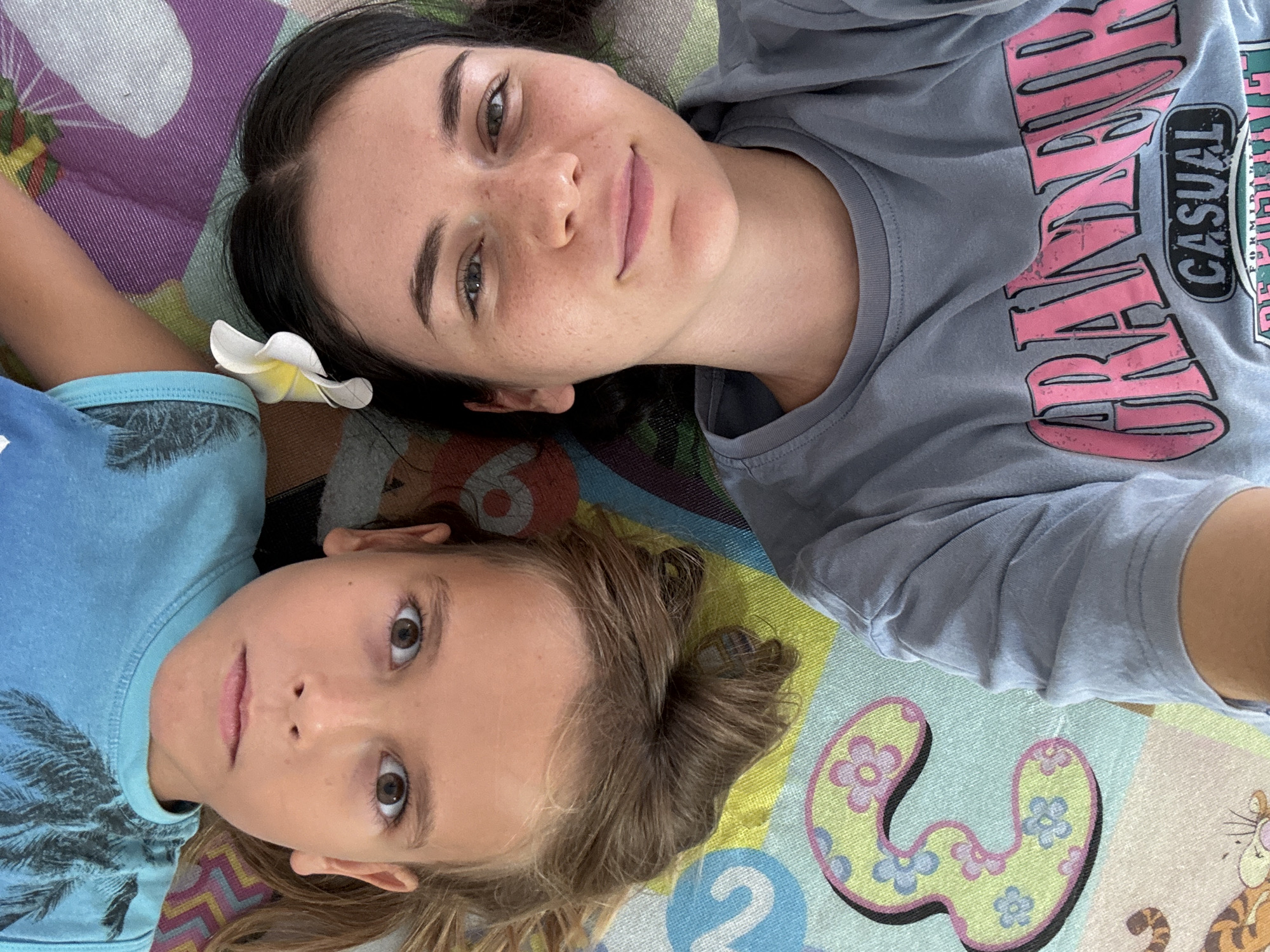 Two individuals, an adult and a child, are lying down on a colorful play mat, likely indoors. The mat features numbers and cartoon animal illustrations. The adult has dark hair, fair skin, and is wearing a grey t-shirt with pink lettering that reads "BRAND NEW CASUAL." The child has light brown hair and is wearing a blue t-shirt with a palm tree graphic. A white flower is placed near the child's ear. The location is likely Mũi Né, Vietnam.