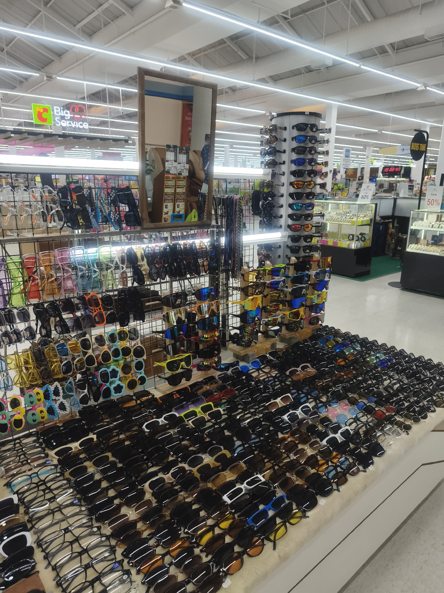 The image shows a display of sunglasses in a large retail store. There are multiple racks and tables filled with sunglasses of various styles, colors, and designs. The store has bright overhead lighting. In the background, there are additional retail displays and signage, including "Big Service" and a "Boss Time" sign indicating the location is in a major store. The "Sale 50%" signs suggest a promotion is running. Based on the presence of the "Big Service" sign, the location is likely a Big C store, suggesting the location is in or around บ้านบางรักษ์, Thailand. There are no people visible in close proximity to the sunglasses display. There are also displays of watches and jewelry in the background.