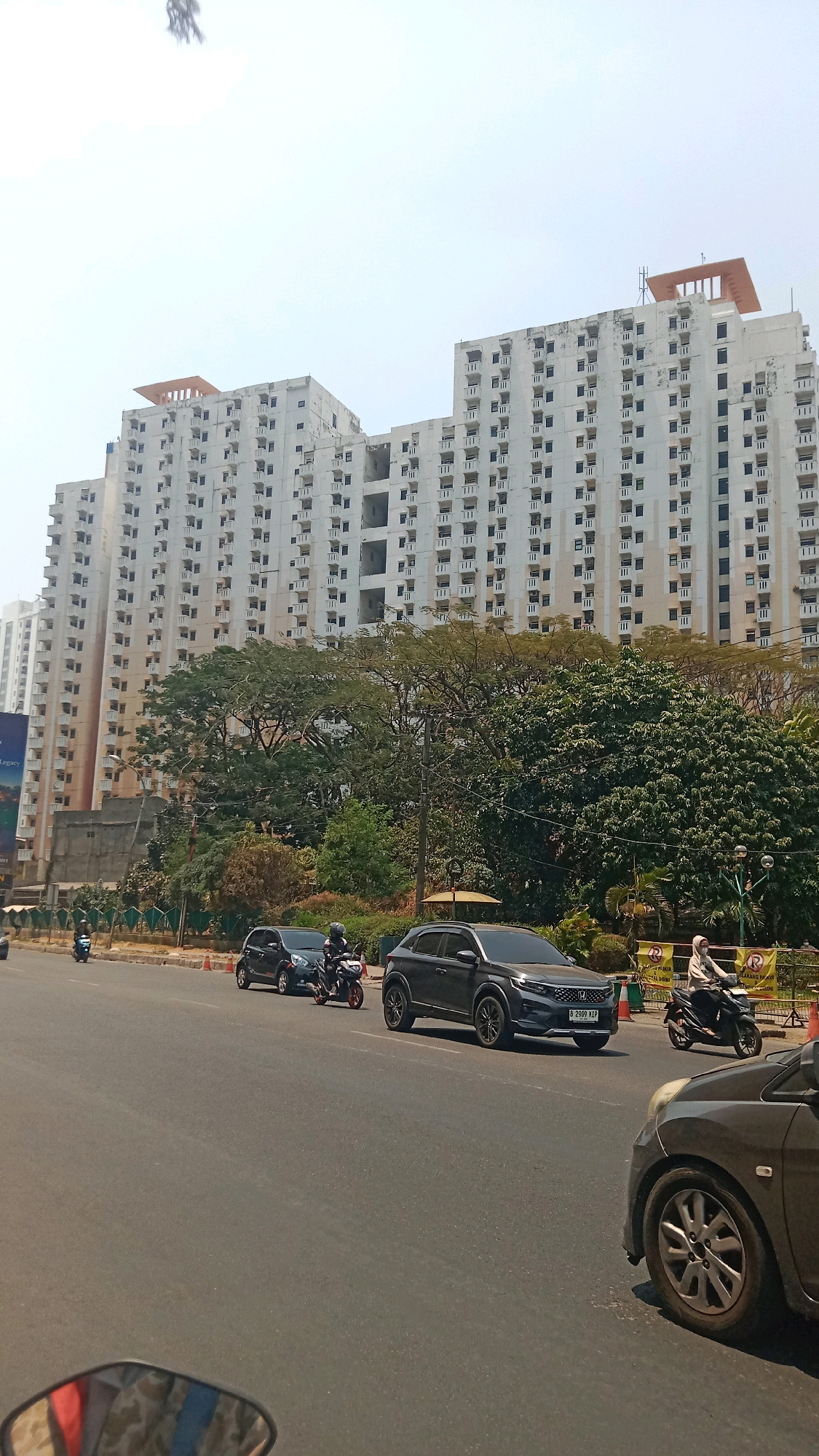 The image captures a street scene in Bekasi, Indonesia, on a clear, sunny day. Dominating the background are two towering, multi-story apartment buildings, their facades covered in numerous small windows and balconies. One building has an unfinished orange structure on its roof, suggesting ongoing construction or renovation. In front of the buildings, lush green trees provide a vibrant contrast to the concrete structures.
The foreground depicts a busy road with moderate traffic. Several vehicles are visible, including a dark gray SUV in the center lane, a smaller dark car to its left, and a motorcycle with a rider in the left lane. Further to the right, another motorcycle with a rider wearing a hijab is also on the road. The street is paved and appears to be a multi-lane road. The overall impression is a typical urban environment in Bekasi, showcasing residential buildings, natural elements, and daily life on the road.