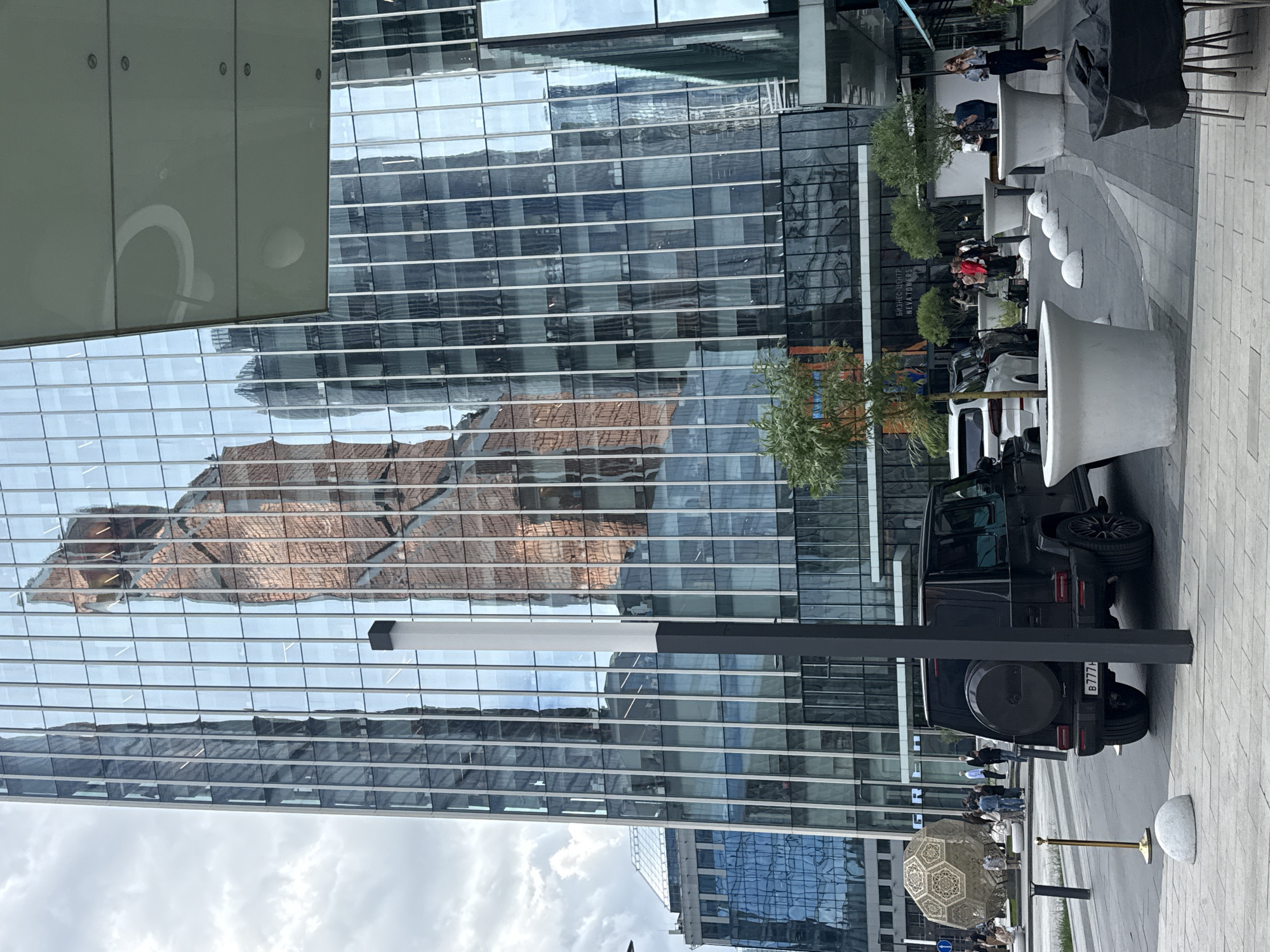 The image depicts an outdoor scene in Khimki, Russia, characterized by modern architecture and urban activity. The focal point is a sleek, black Mercedes-Benz G-Class SUV parked on a paved walkway. The background showcases contemporary buildings with glass facades that reflect the surrounding environment, including an older brick building. Several people are present, some standing near a decorative golden umbrella, while others are seated at outdoor tables, some of which are shaded by trees. Architectural elements like large, white planters and minimalist streetlights contribute to the modern aesthetic. The overall impression is one of a bustling urban environment with a blend of modern and classic architectural styles.