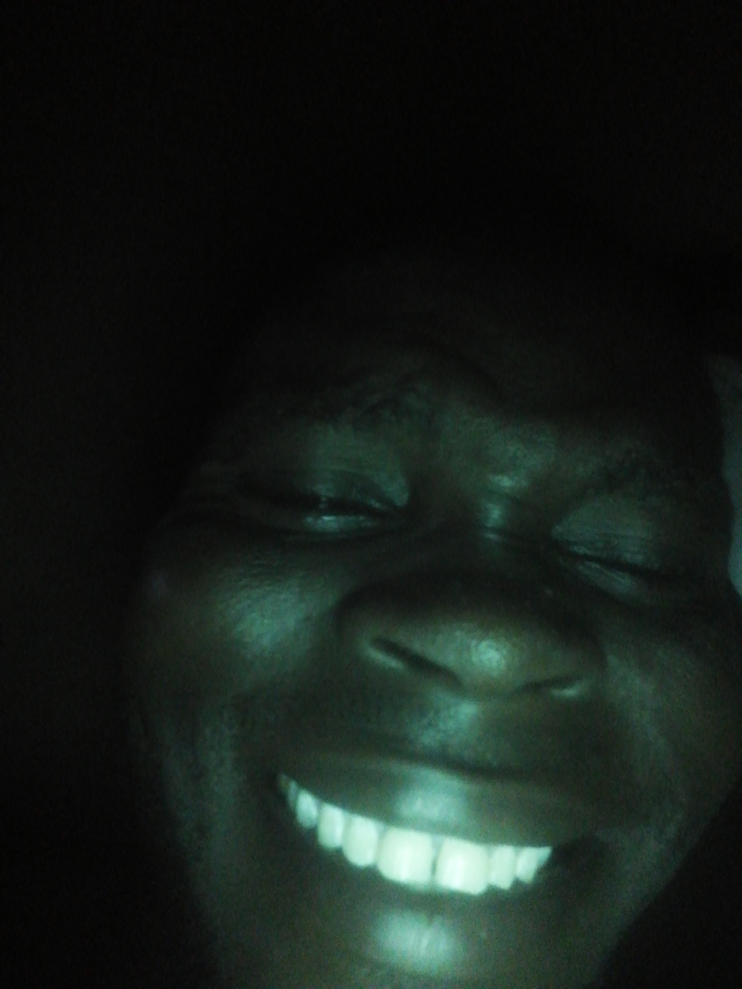 A close-up image of a person's face, illuminated with a green-tinged light. The person has dark skin and is smiling, revealing white teeth. The eyes are closed. The background is dark and indistinct. The location is Unknown, Unknown.