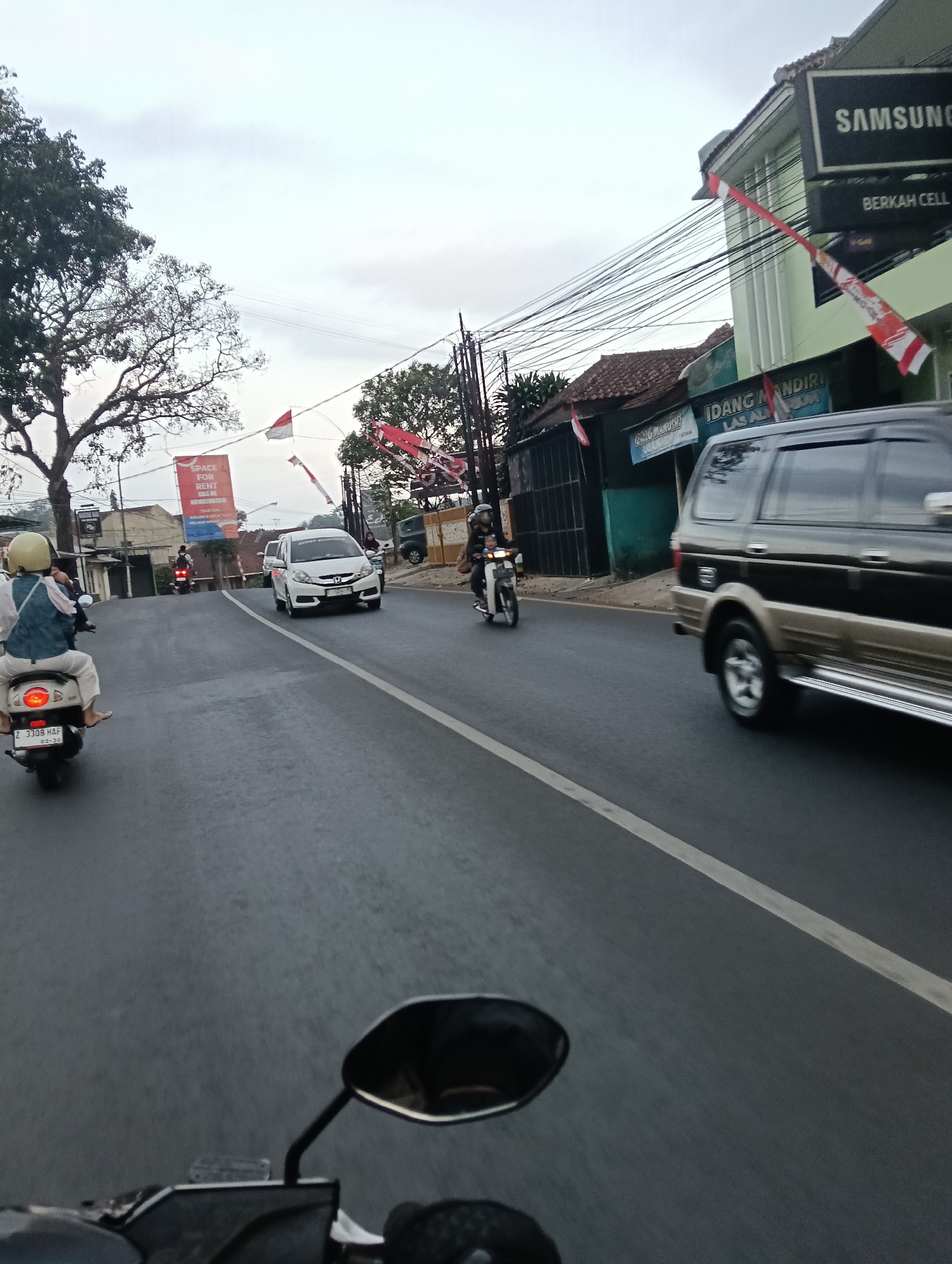 The image depicts a street scene in Tasikmalaya, Indonesia, likely during the late afternoon or early evening. The foreground is dominated by the perspective of a person riding a motorcycle, with a rearview mirror and handlebars visible. The road surface is asphalt, marked with a white line.
Several vehicles are on the road. A white car, possibly a Honda Mobilio, is traveling in the opposite direction. A dark-colored SUV, possibly a Mitsubishi Pajero Sport, is also in view. Multiple motorcycles are present, including one in the middle ground with a rider wearing a white helmet and a blue shirt, and another further ahead. The rider of the motorcycle in the foreground is wearing a white helmet and a blue and white garment.
The environment includes buildings lining the street. One building has a large "SAMSUNG" sign and another sign that reads "BERKAH CELL". Another building displays a banner with "SPACE FOR RENT". Red and white Indonesian flags are visible, hanging from poles and buildings, suggesting a national celebration or event. Trees with sparse foliage are also part of the scenery. Power lines are strung across the scene, contributing to the urban landscape. The sky is overcast, indicating cloudy weather. The overall impression is a moderately busy street in an Indonesian city.