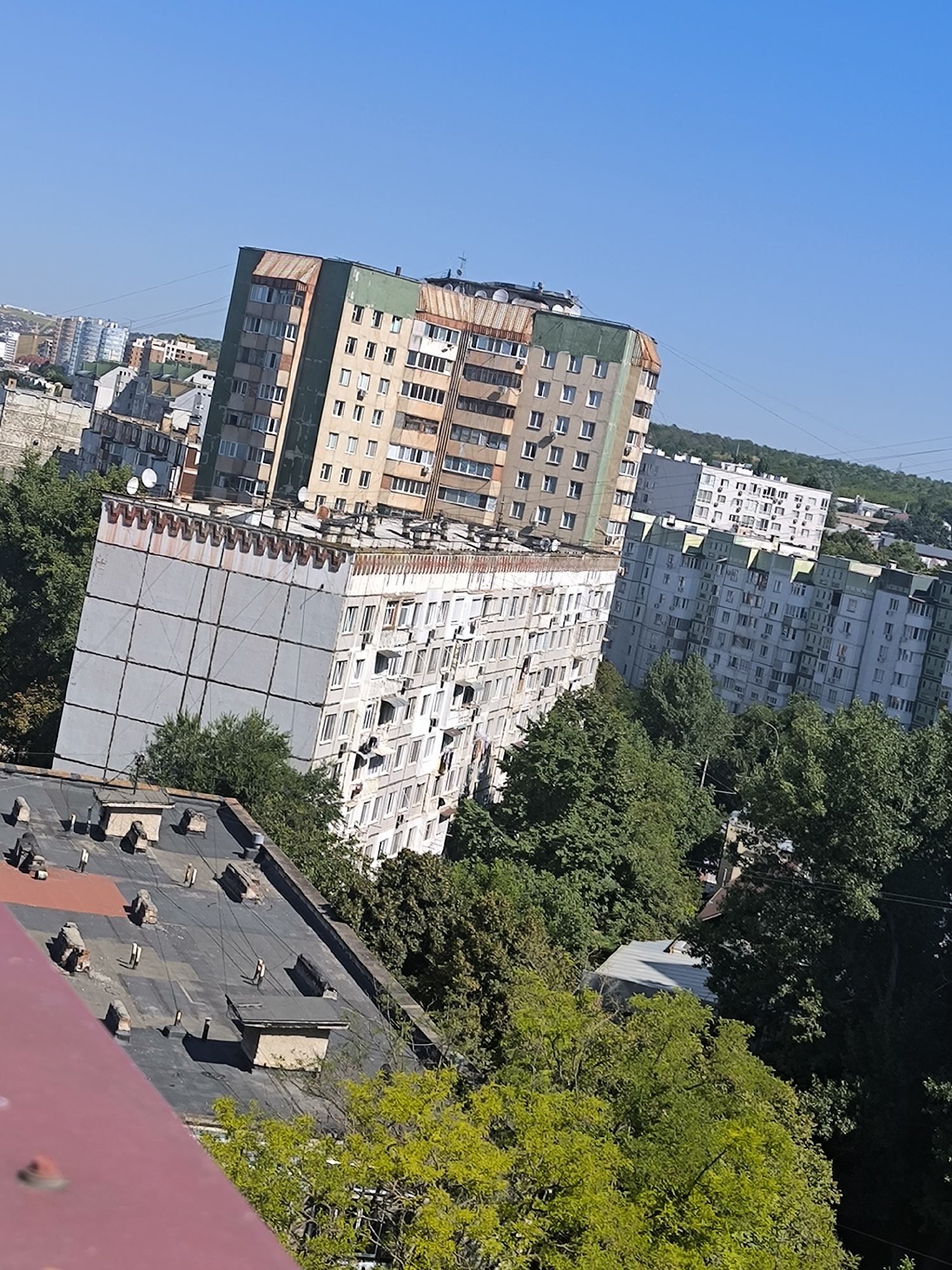 The image displays a collection of Soviet-era apartment buildings against a clear blue sky. The buildings are characterized by their multi-story design, repetitive window patterns, and a somewhat weathered appearance. In the foreground, the roof of a lower building is visible, showing a dark surface with various rooftop structures and some debris. Beyond this, several taller apartment blocks dominate the frame, with one particularly prominent building featuring a green and brown facade. Trees, some with lush green foliage and others with lighter green leaves, are interspersed among the buildings, suggesting a residential area with some greenery. The overall impression is one of an urban landscape, likely in Eastern Europe, given the architectural style. The image seems to have been taken from an elevated position, looking down on the cityscape. Without specific landmarks, it is impossible to pinpoint the exact location within Chișinău, Moldova, but the architectural style is consistent with that found in many post-Soviet cities.